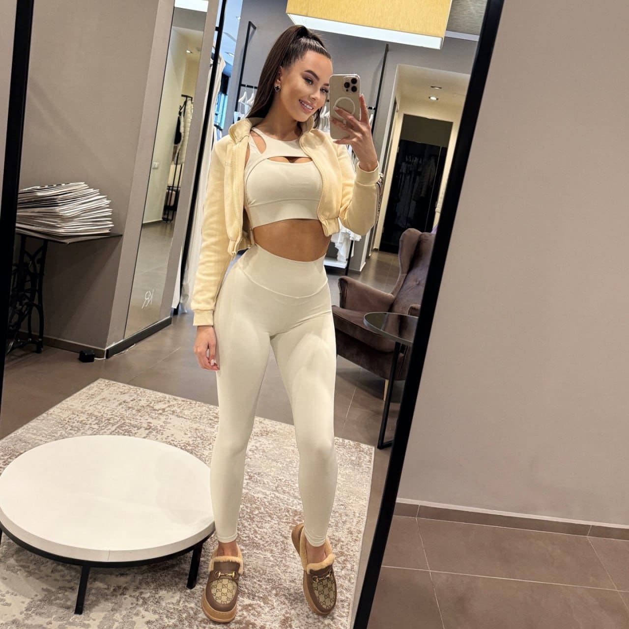 A woman in cream cut-out activewear and fur-trimmed loafers stands in a fitting room, taking a mirror selfie near a round table.