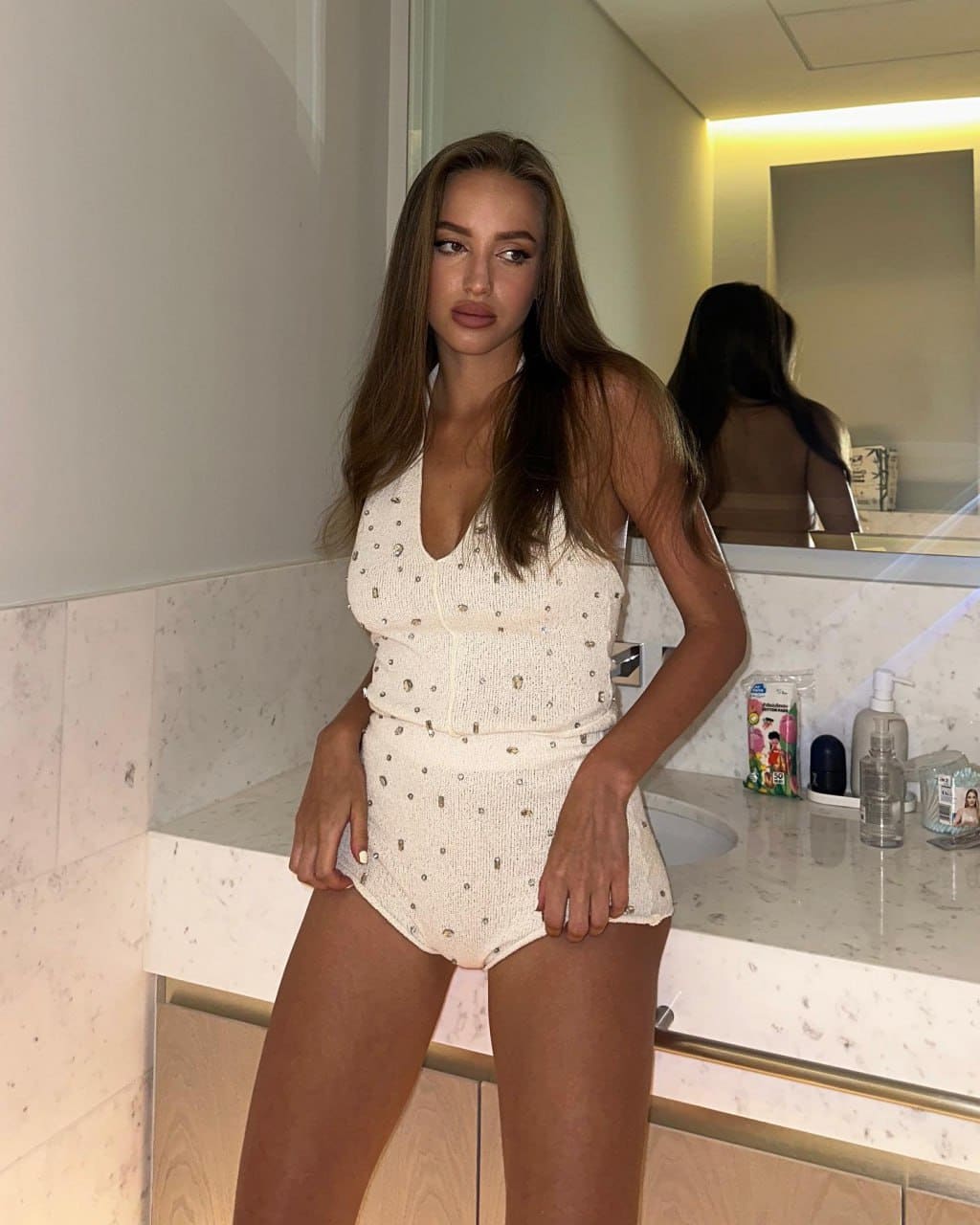 A woman in a white studded swimsuit poses with hands on hips in front of a marble bathroom countertop and mirror.