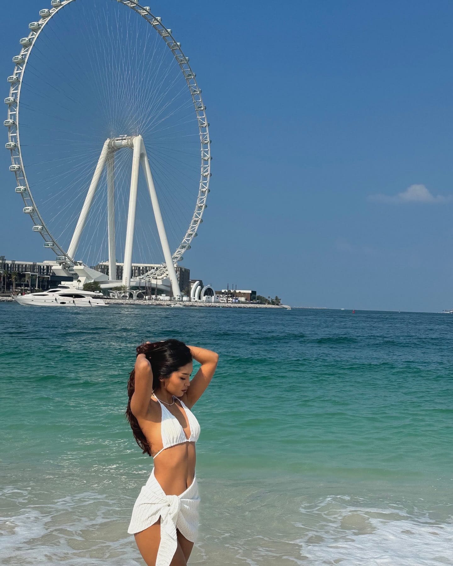 A woman in a white bikini and wrap skirt stands in seawater with an observation wheel in the background.