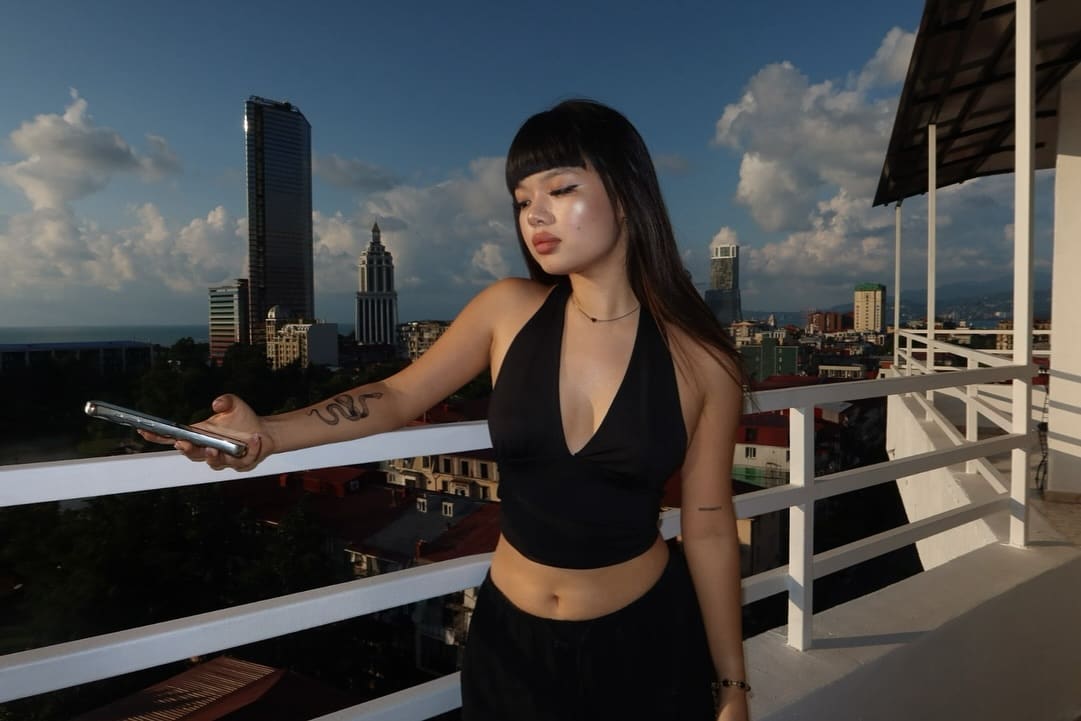 A woman in a black halter top and pants stands on a balcony holding a smartphone, facing forward against a city skyline.