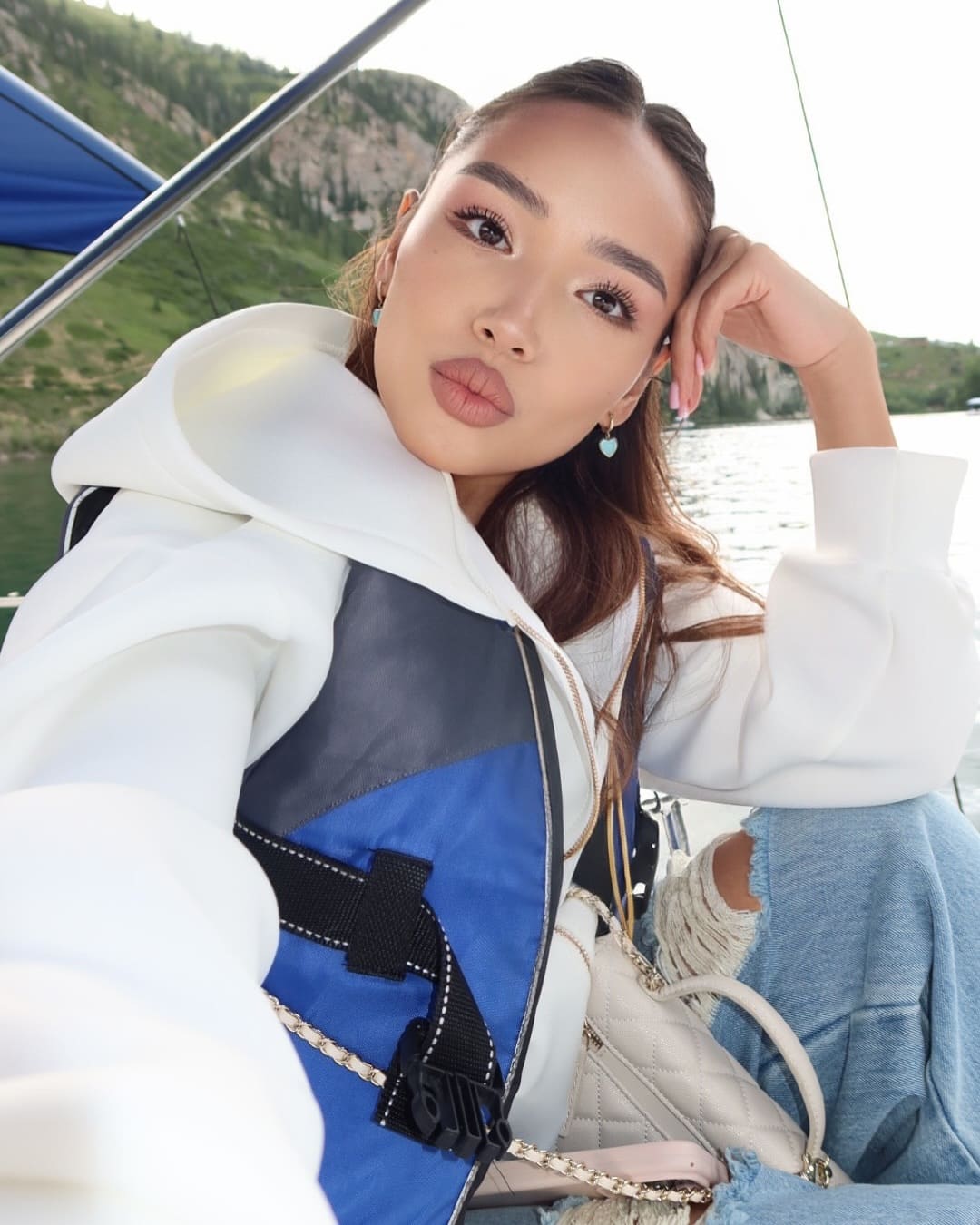 Mirelia sits on a boat in a white hooded jacket and life vest, hand on head, with a quilted bag on her lap against water and hills.