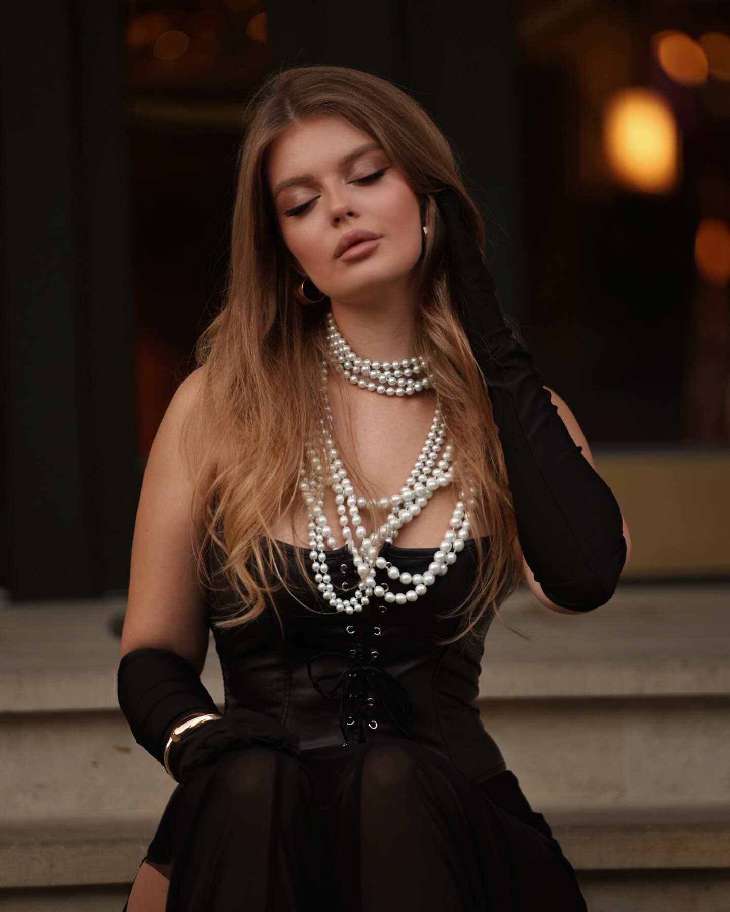 A woman sits on stone steps in a black lace-up corset, sheer skirt, and gloves, wearing white pearls with one hand raised to her hair in dim light.