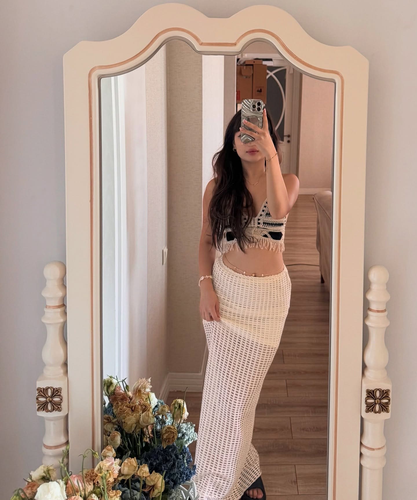 A woman in a crocheted crop top and open-weave skirt takes a mirror selfie, dried flowers on the floor beside her.