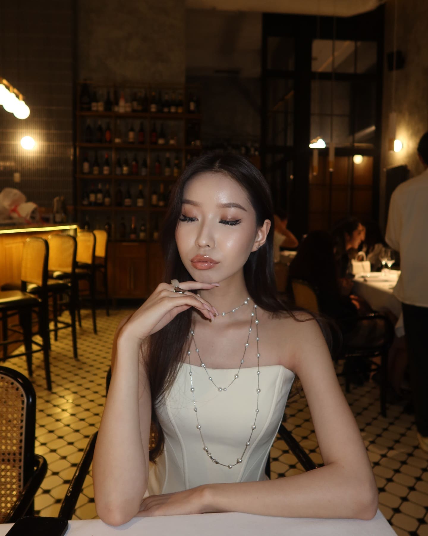 A woman sits at a dimly lit restaurant table in a cream strapless top and beaded necklace, resting her hand near her chin.