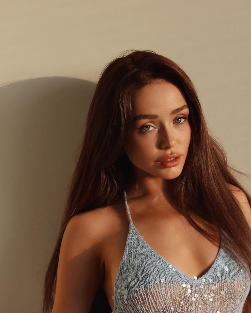 Grace sits in a chair wearing a light blue sequined mesh top with thin straps against a plain beige wall.