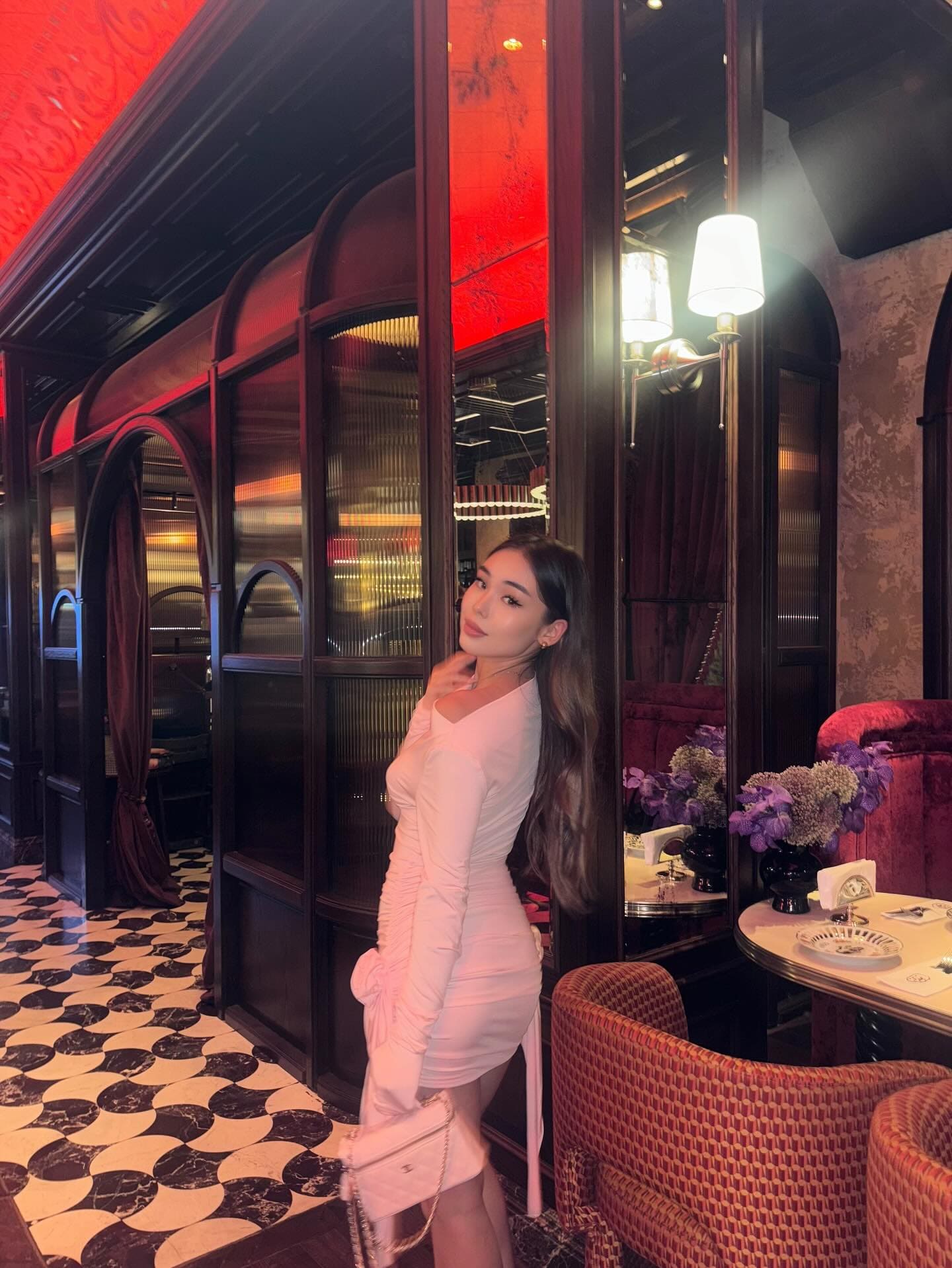 A woman stands in a restaurant with red walls and checkered floors, wearing a pink dress and holding a small white handbag.