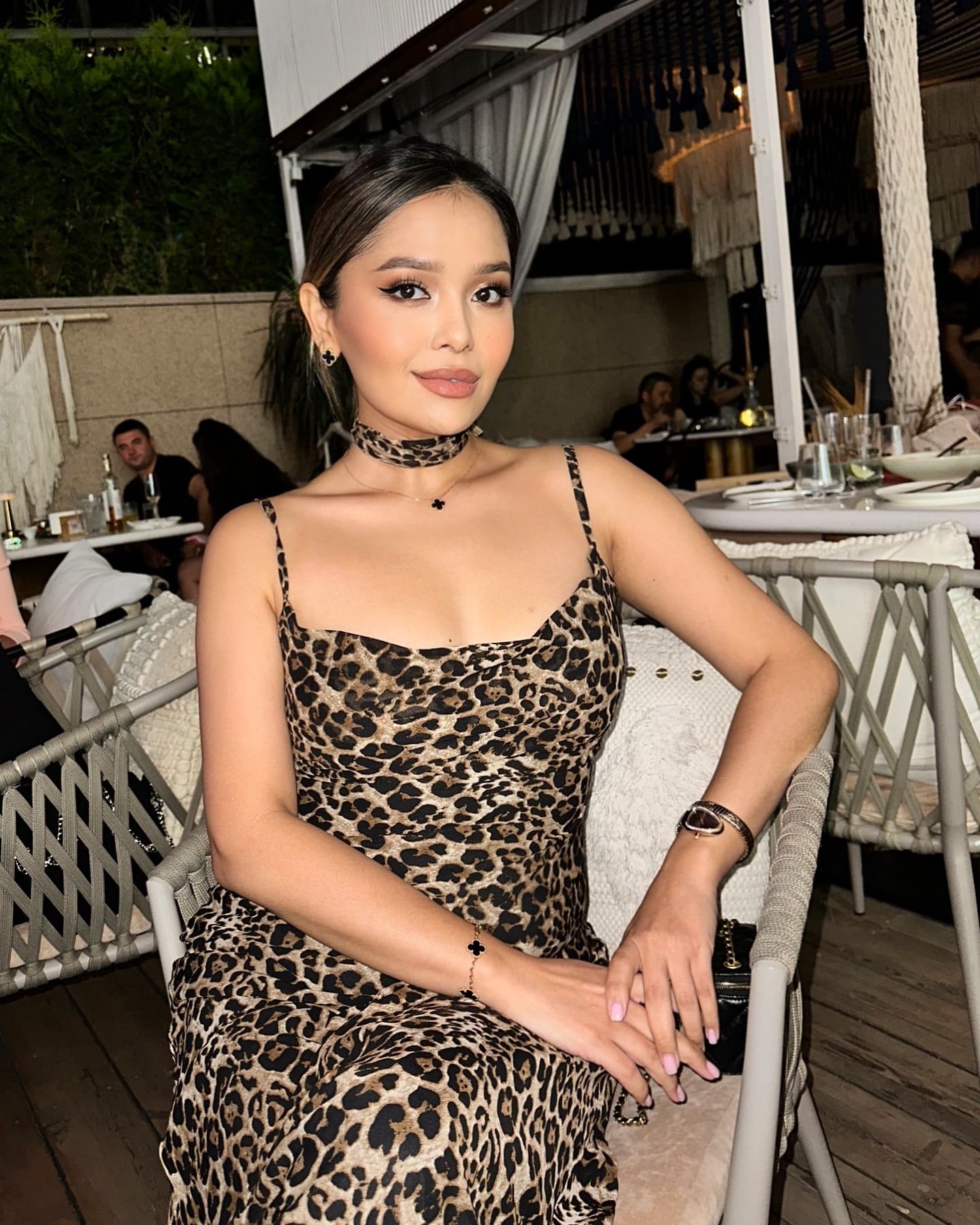 Aknur sits in a woven chair at an outdoor restaurant, wearing a leopard-print dress and choker, with hands resting on her lap.