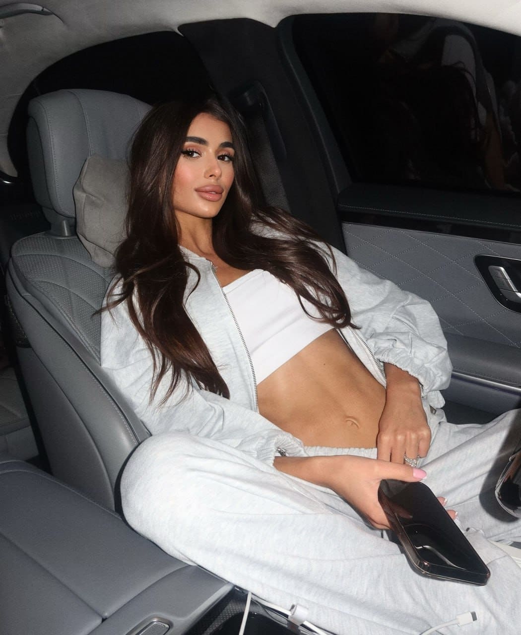 A woman sits in a car back seat wearing a grey hoodie over a white crop top and sweatpants, holding a phone.