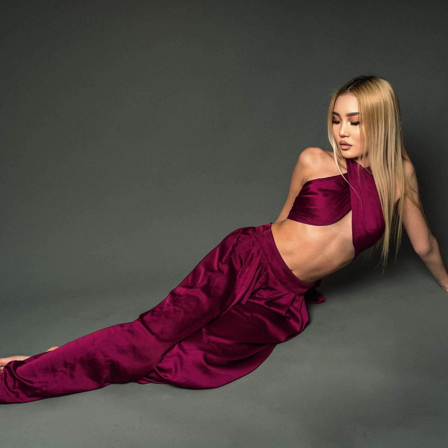 A woman lies on her side on a gray studio floor, wearing a one-shoulder burgundy satin crop top and matching high-waisted satin trousers, with her head turned toward the camera.