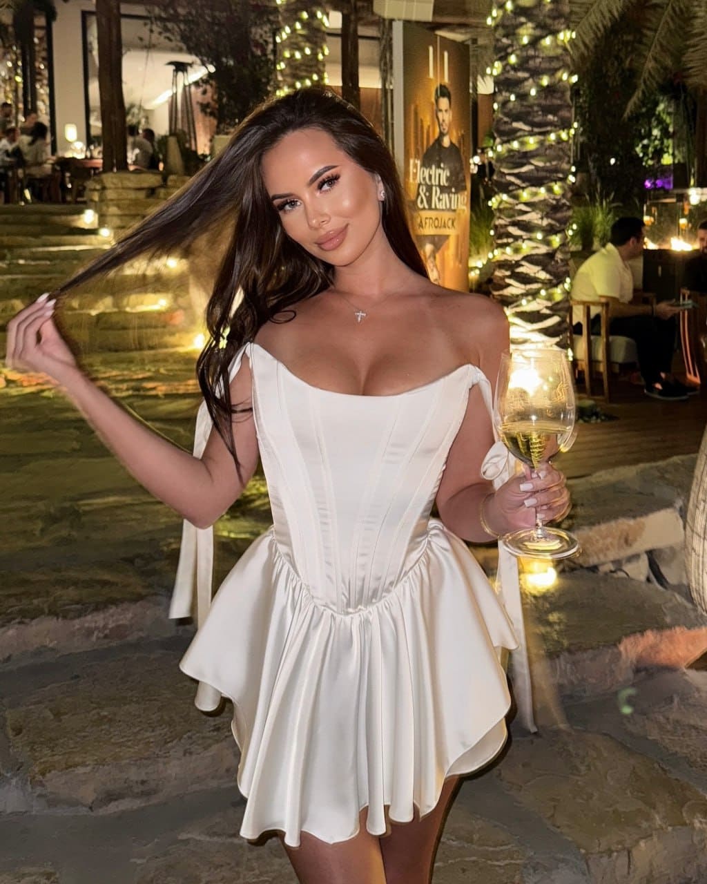 A woman in a white off-shoulder mini dress holds wine outdoors at night, with string-lit trees and a patio in the background.