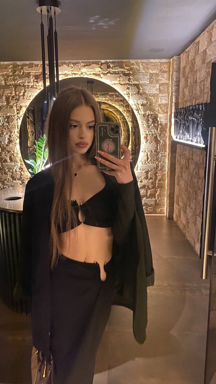 A woman takes a mirror selfie in a stone room, wearing a black bralette, low-slung skirt, and open blazer while holding a phone.