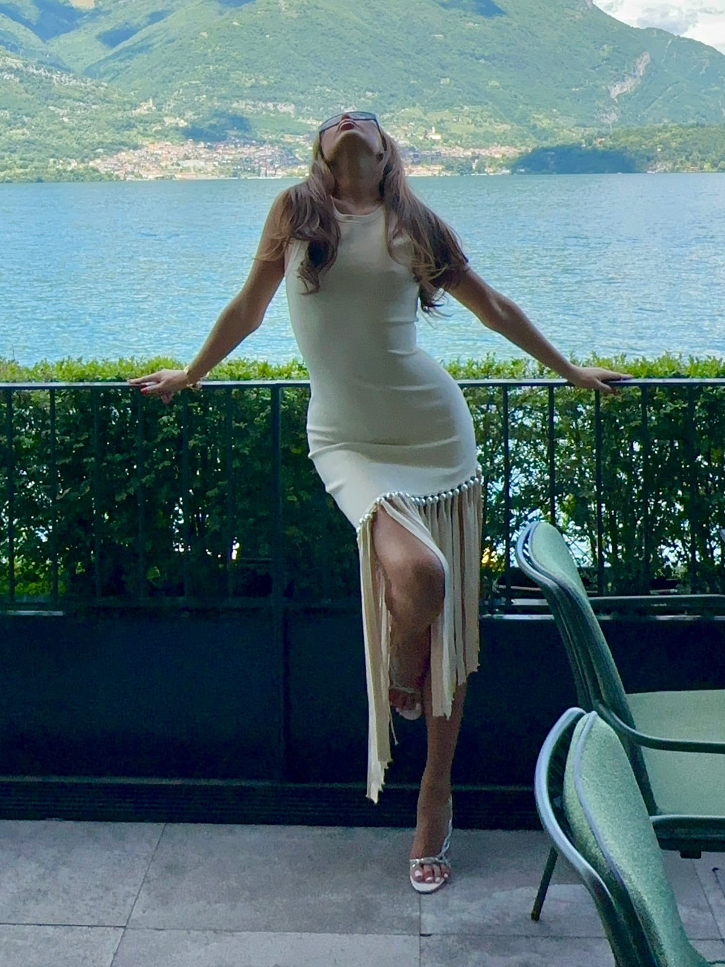 A woman stands on a terrace in a cream sleeveless dress with a high slit and fringe hem, leaning on a railing with a lake and hills behind.