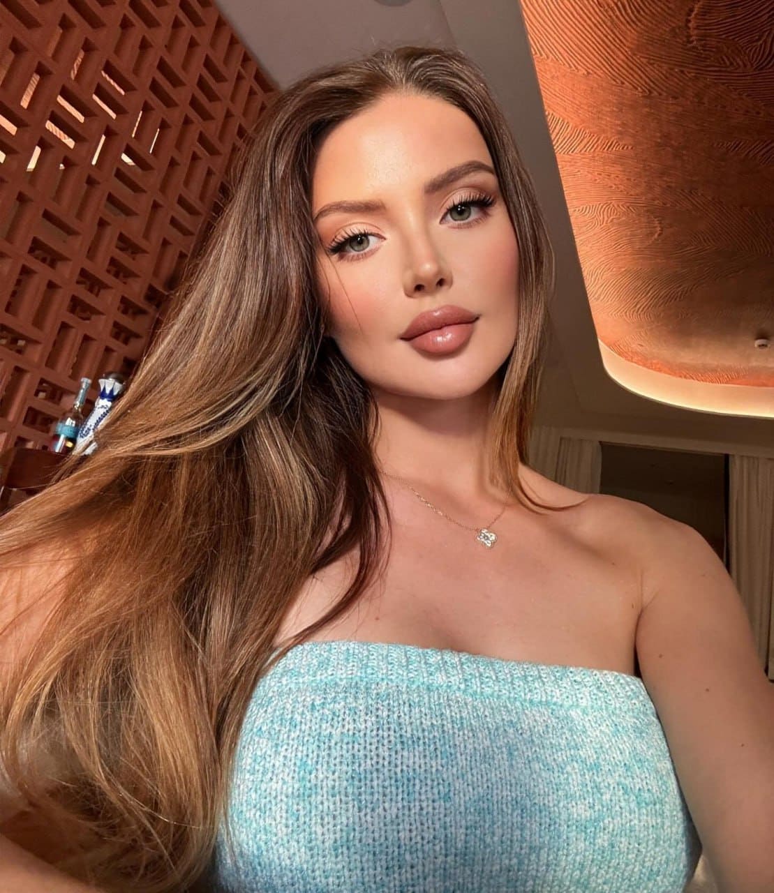 Vita in a strapless light blue textured top and necklace, posing indoors against a patterned wooden wall with warm ceiling lighting.
