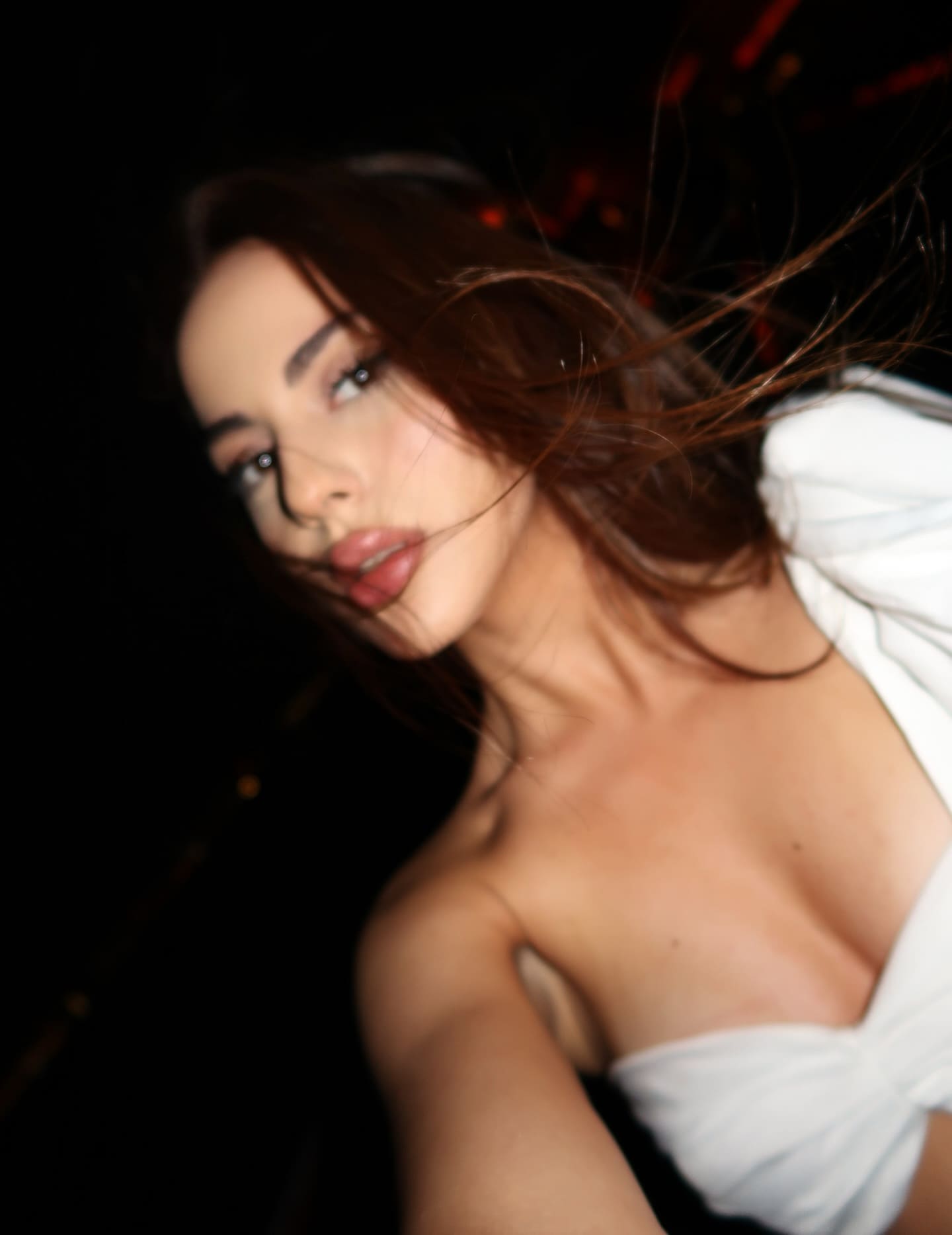A chest-up portrait shows a woman in a white off-the-shoulder garment, hair blowing across her face, against a dark background with red lights.