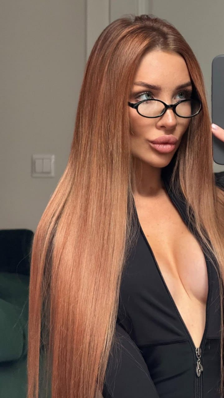 A woman with reddish-brown hair and glasses wears a black V-neck top indoors, holding a phone near a light wall and dark sofa.