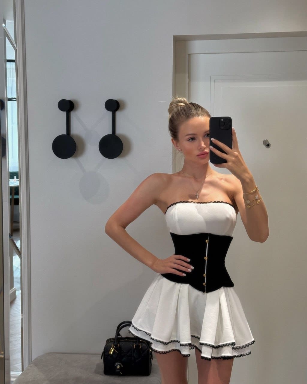 A blonde woman with a bun wears a white strapless top with a black corset waist and pleated skirt, standing near a white door.
