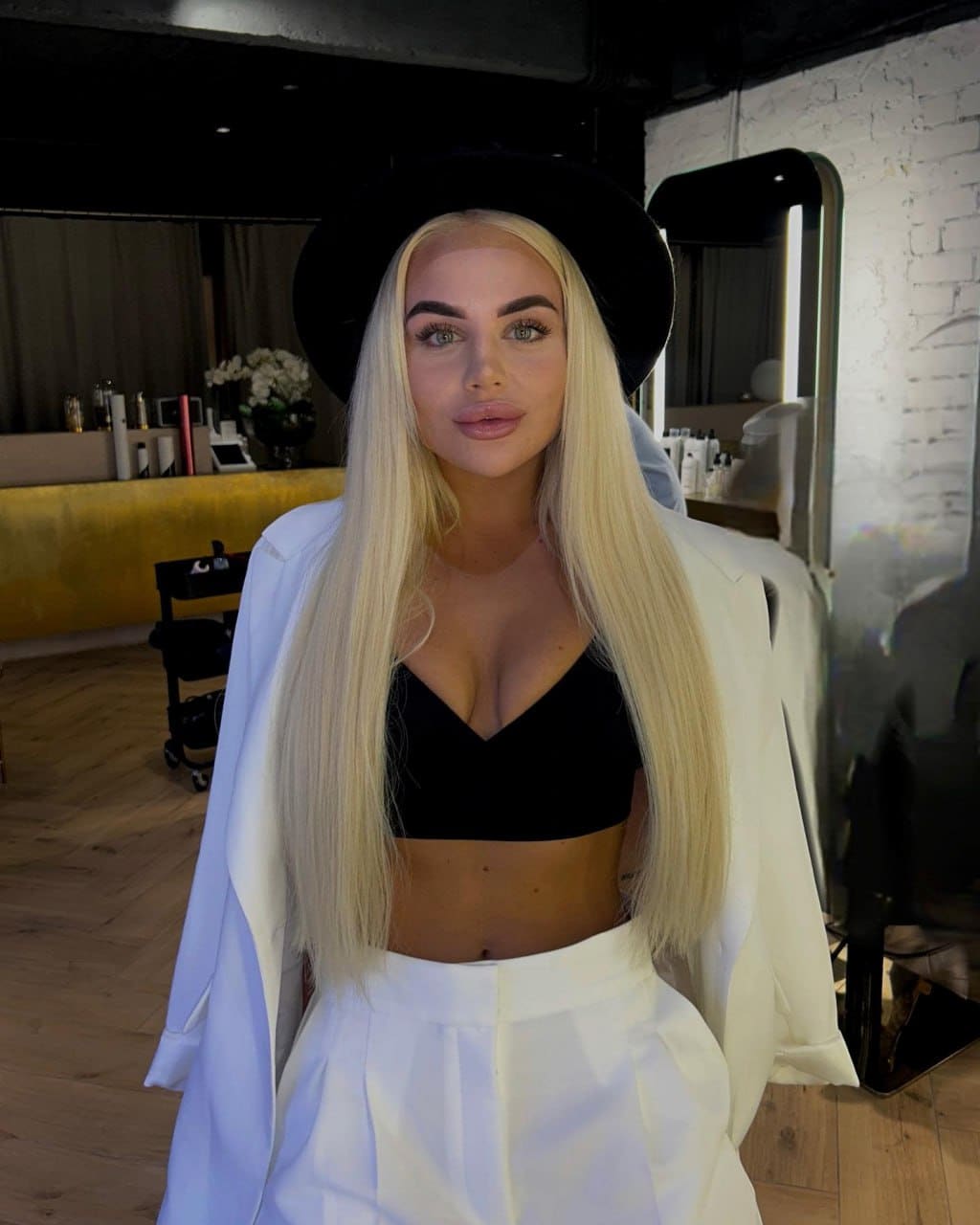 Facing the camera in a white blazer, black bralette, and wide-brimmed hat, standing near a mirror and brick wall.