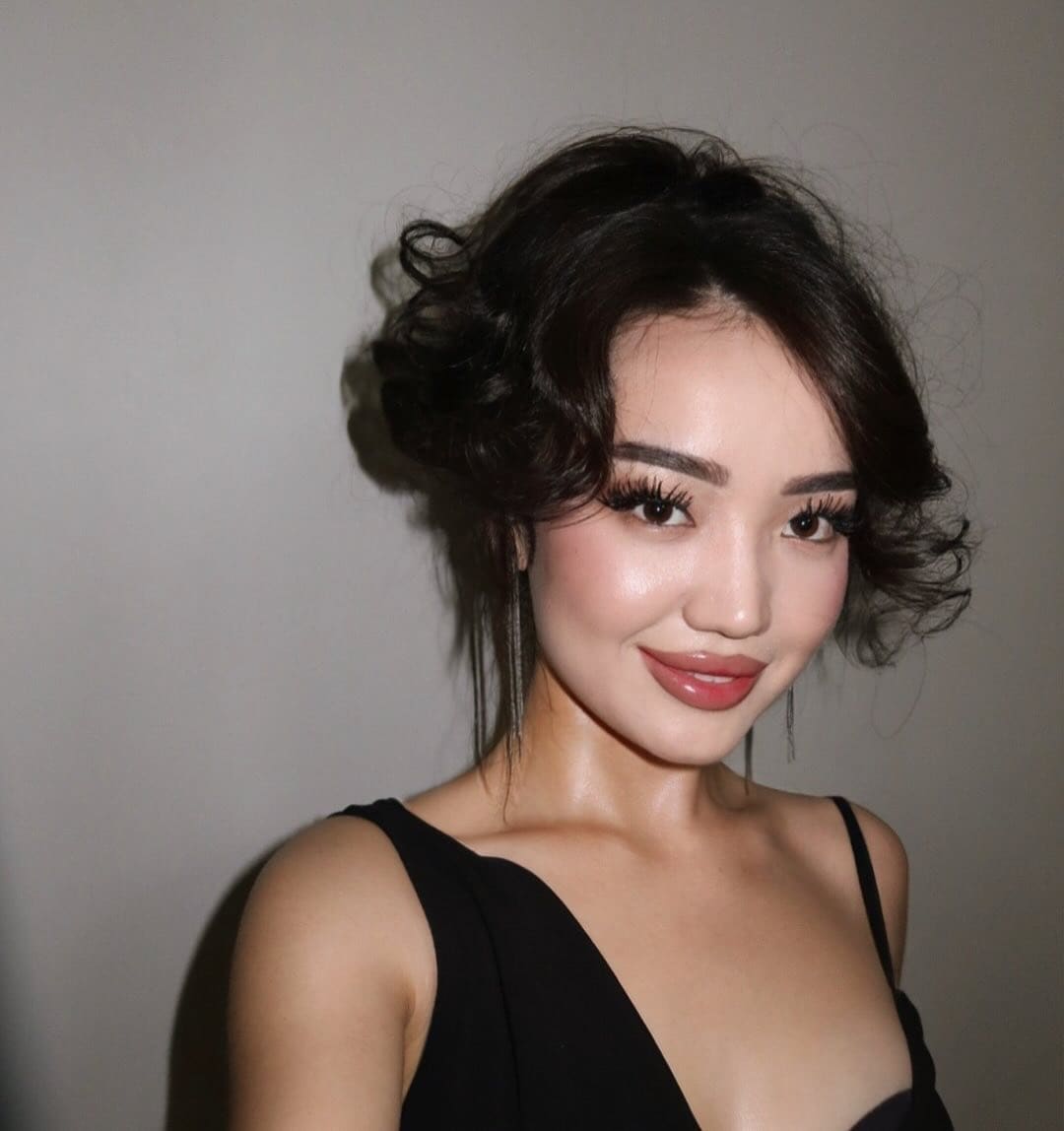 A woman with voluminous curled hair wears a black sleeveless top with a deep V-neckline, posing against a plain light wall.
