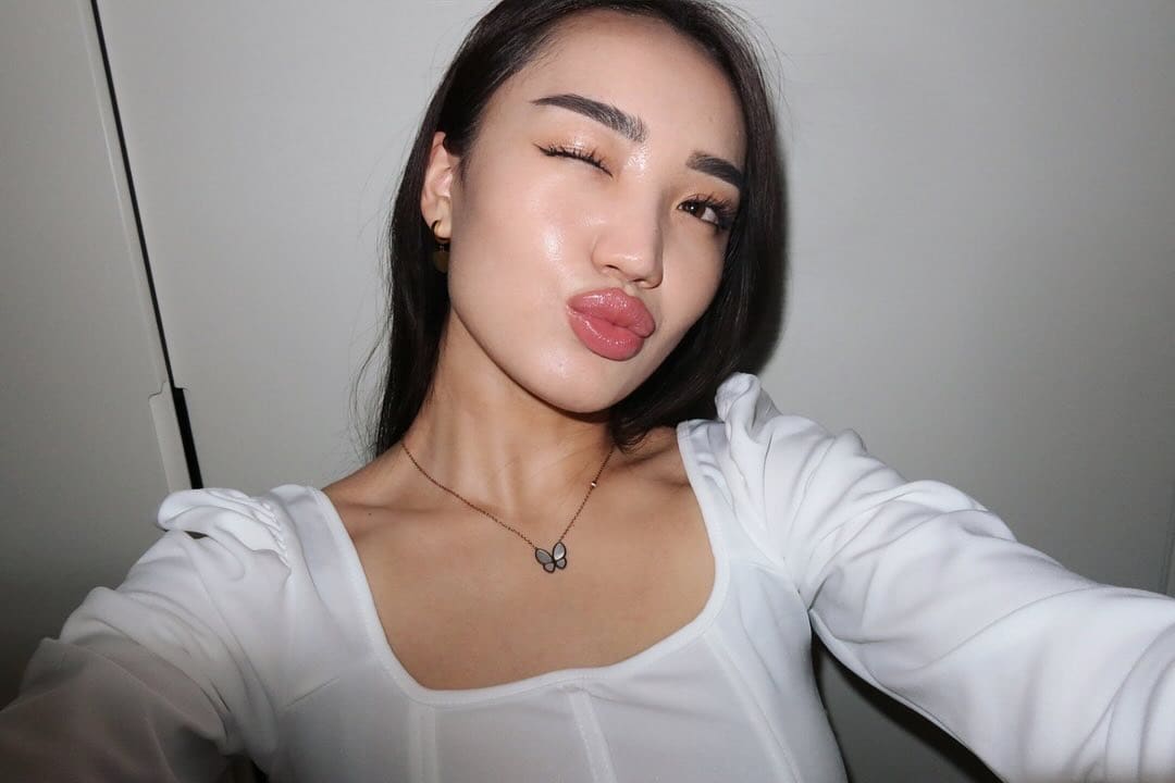 A close-up selfie shows a woman winking in a white long-sleeved top with a square neckline and a butterfly pendant necklace.