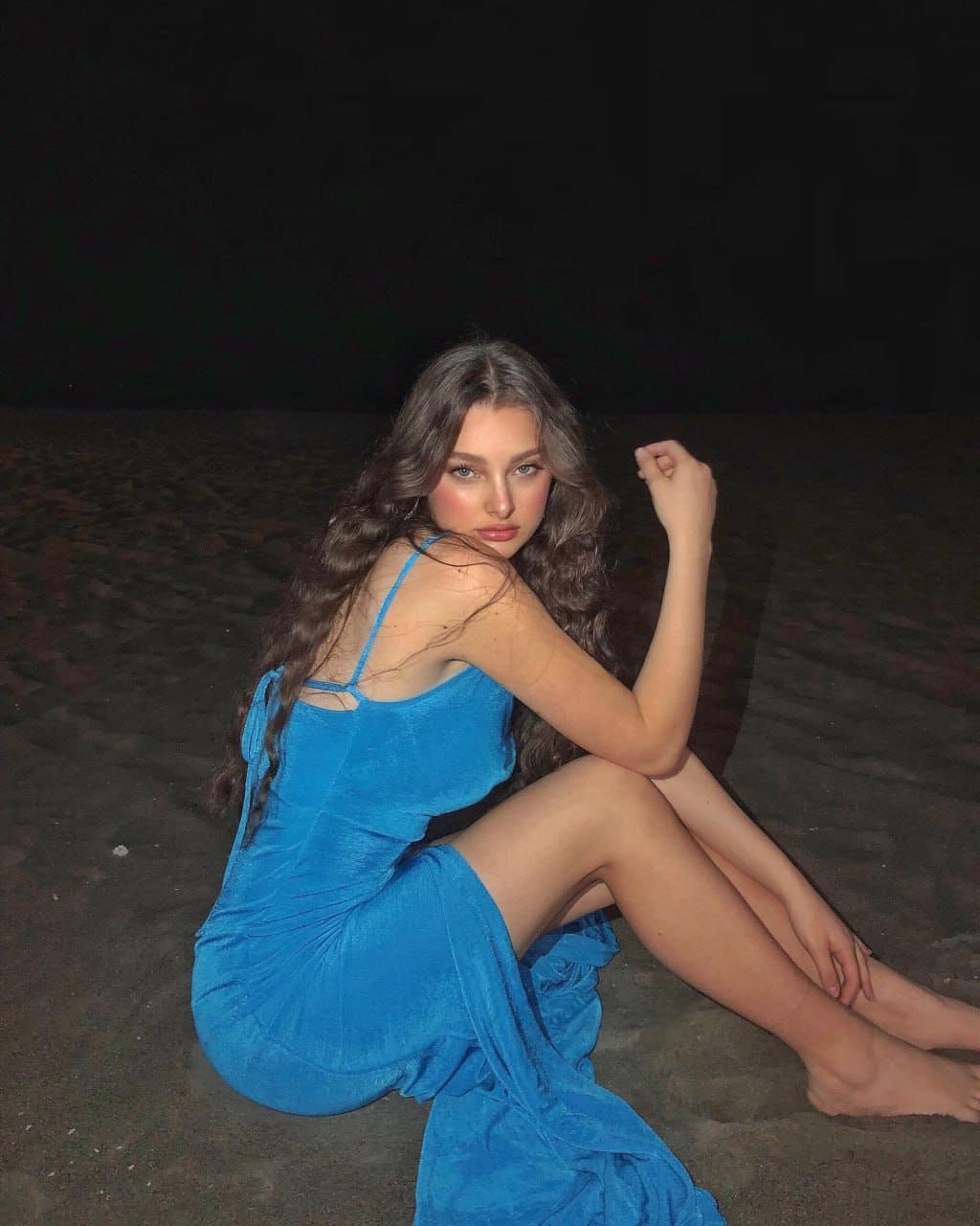 A woman sits barefoot on dark sand at night in a blue dress with a high slit, one arm raised near her shoulder.
