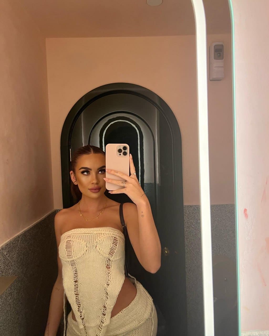 A woman takes a mirror selfie in a hallway wearing a cream open-knit two-piece outfit, holding a phone.
