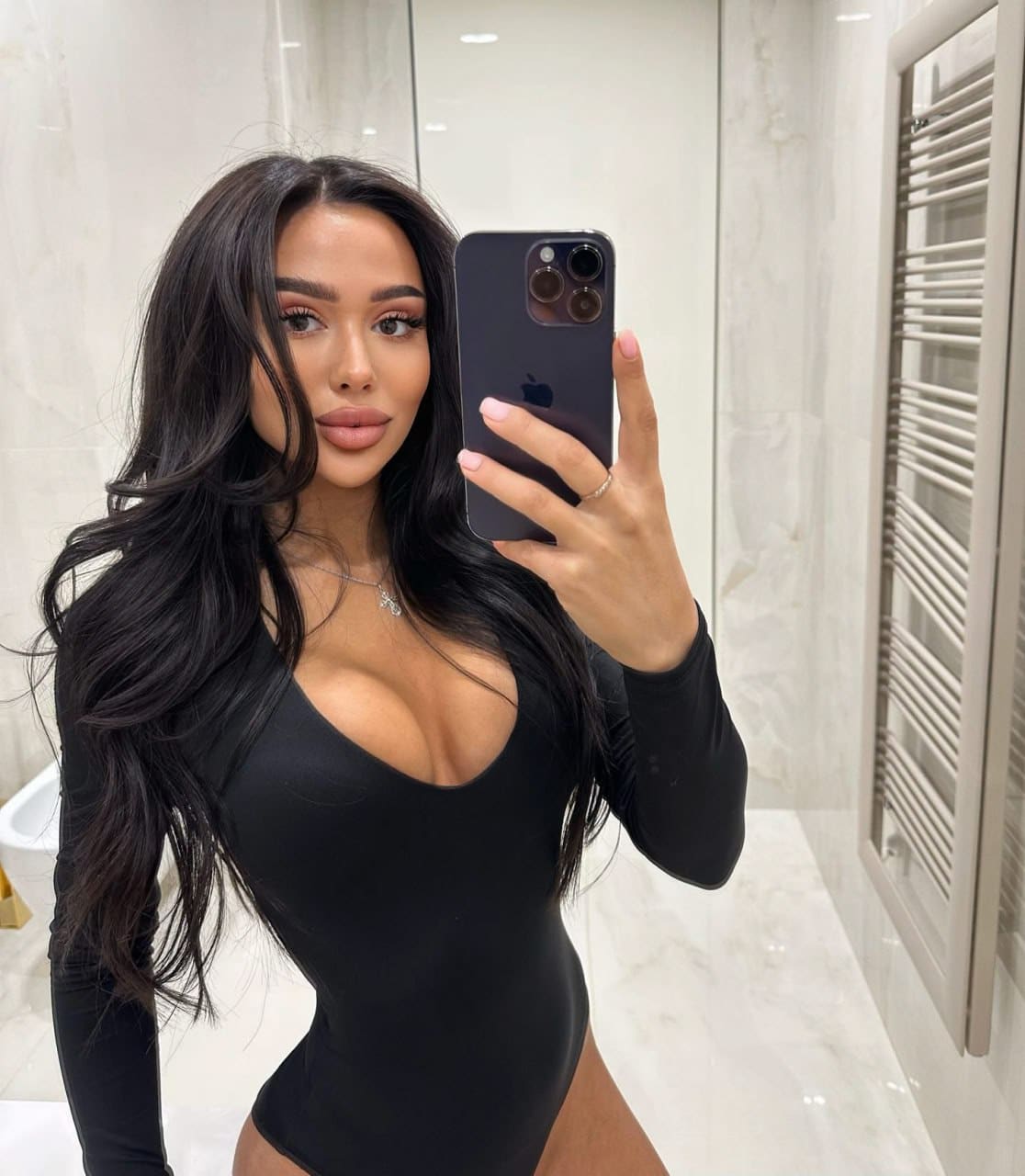Woman in a black long-sleeved bodysuit takes a bathroom mirror selfie, holding a dark smartphone and wearing a thin necklace.