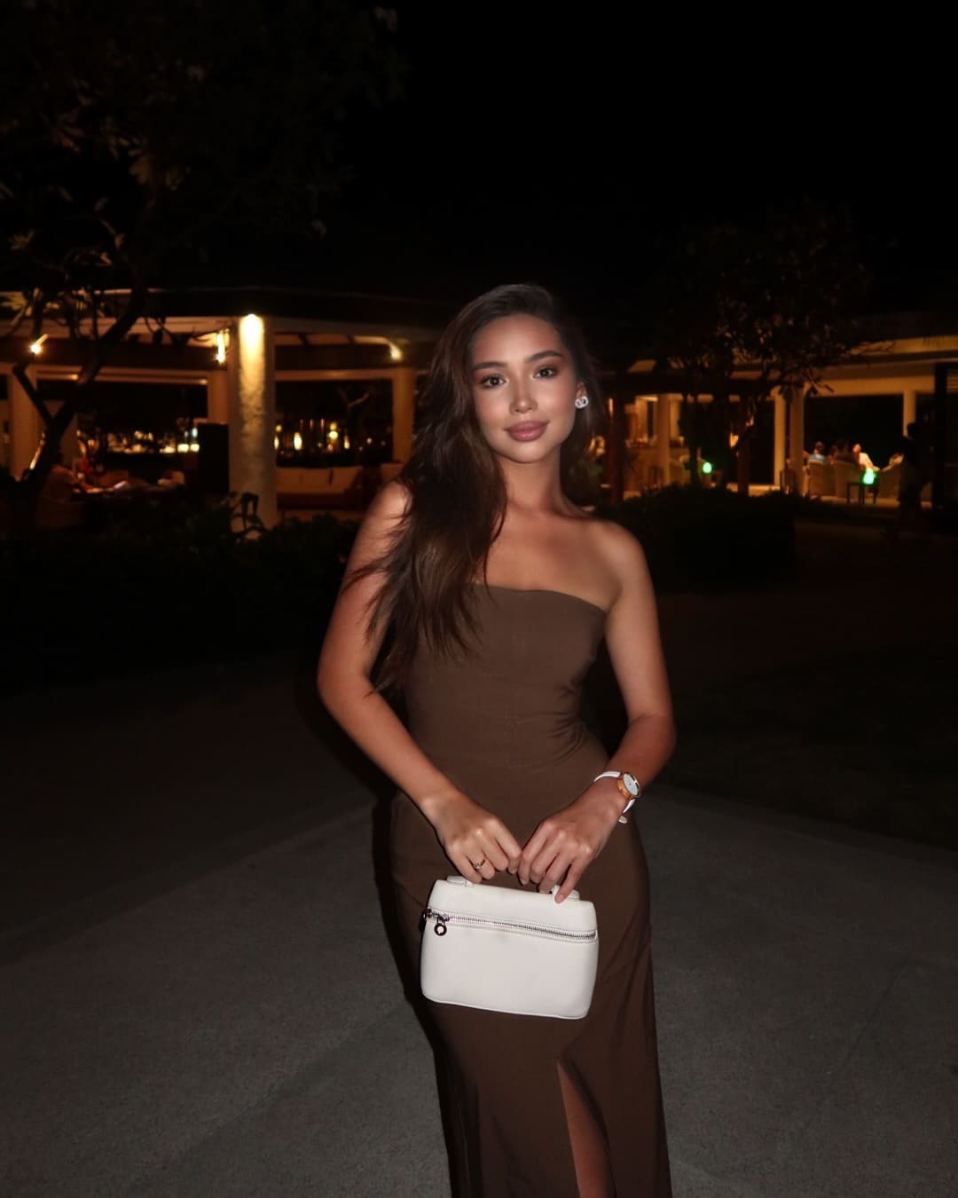 Woman in strapless brown floor-length dress holds a white handbag outdoors at night, with lit buildings and trees behind.