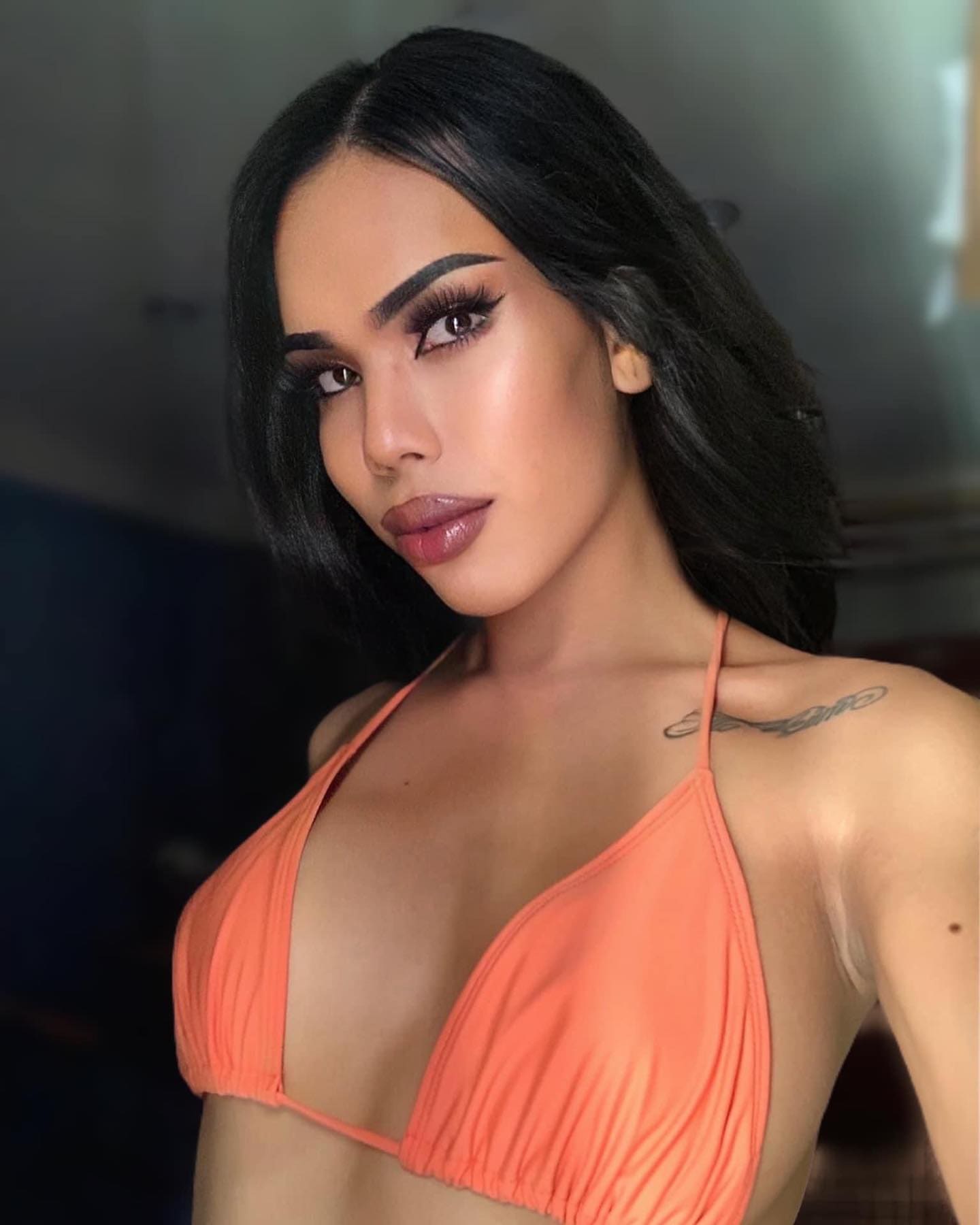 Kamila with long dark hair and a shoulder tattoo wears an orange halter bikini top against a dim indoor background.