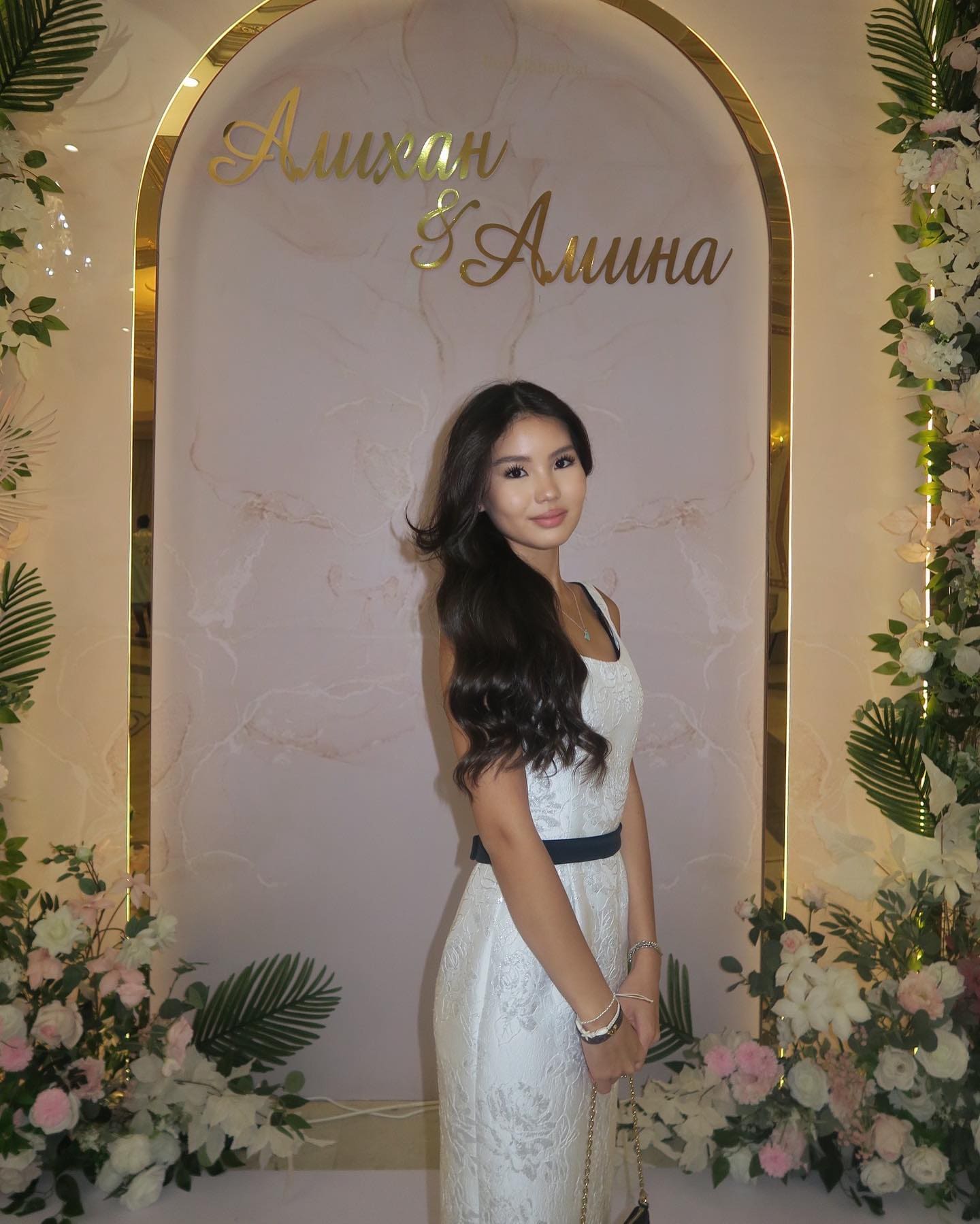 A woman in a white sleeveless lace dress with a dark belt stands before a marble arch decorated with flowers and gold lettering.