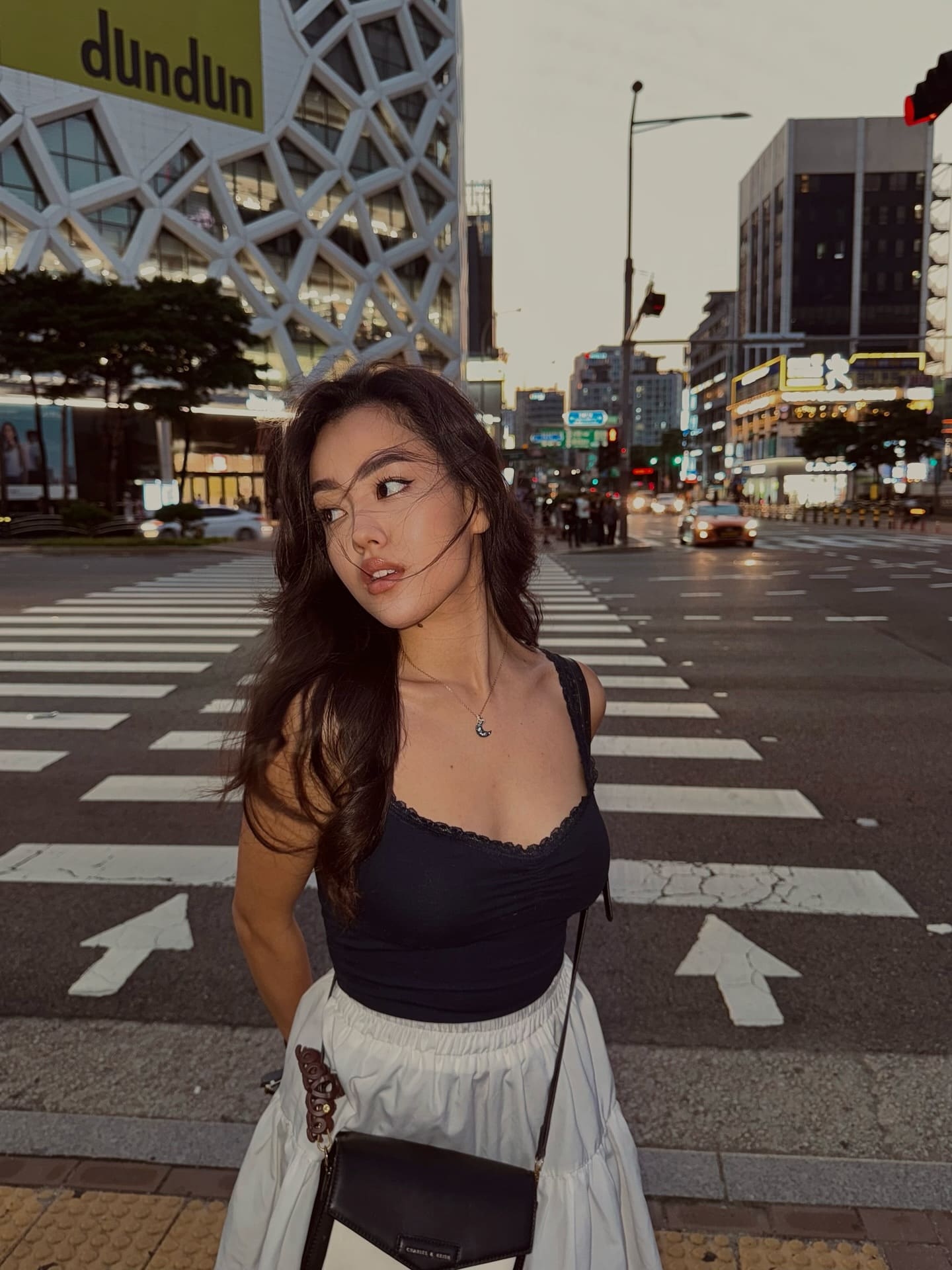 A woman in a black camisole and white skirt stands on a city crosswalk at dusk, looking over her shoulder with a small black crossbody bag.