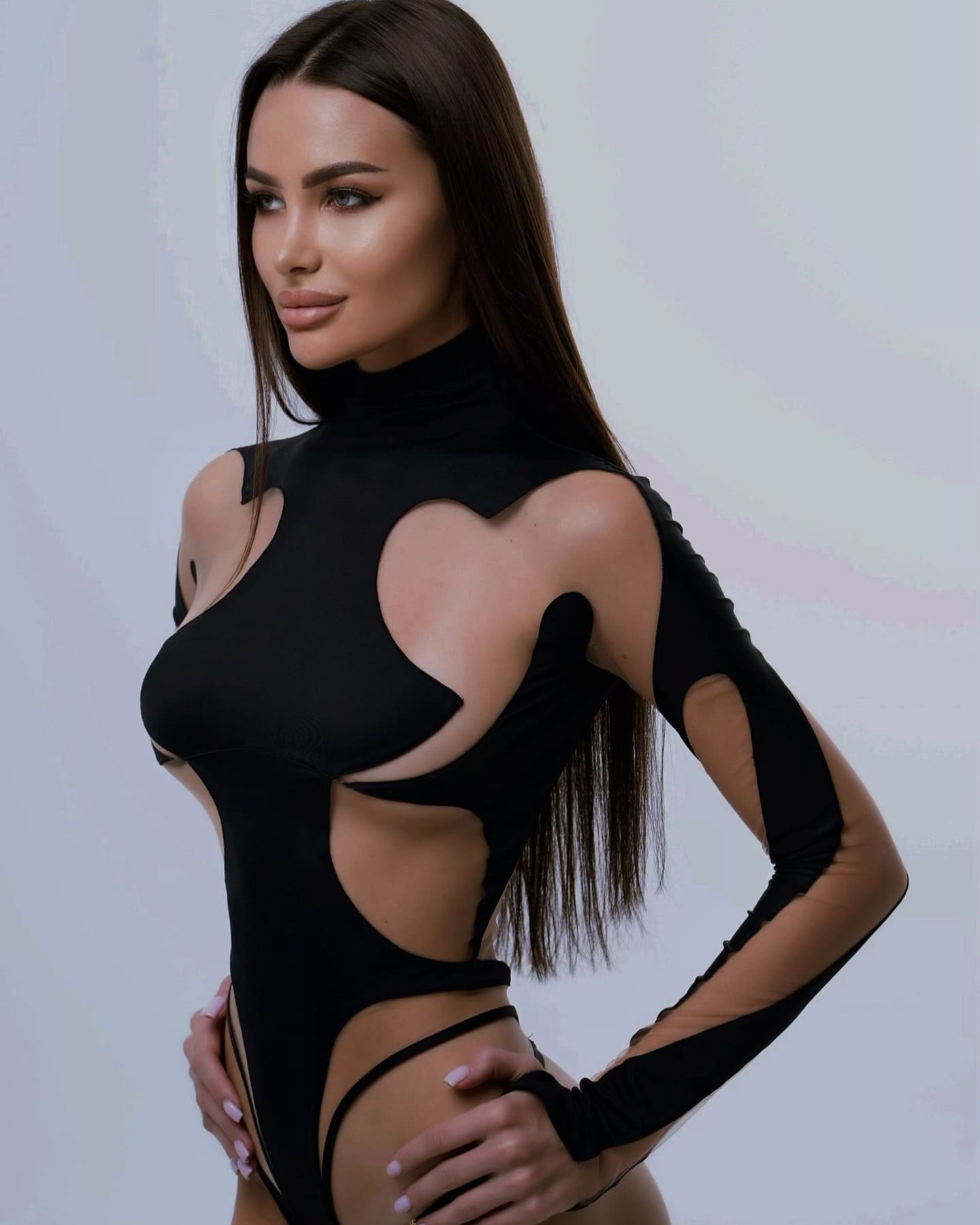 A woman in a three-quarter pose wears a black high-neck bodysuit with large cutouts, set against a plain light background.