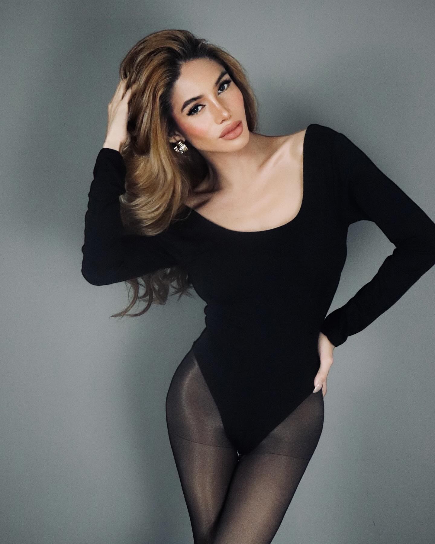 Nancy stands against a gray backdrop in a black bodysuit and sheer tights, one hand on her hip and the other in her hair.