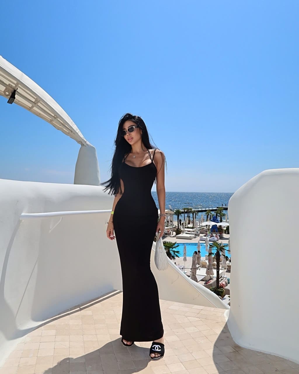 Woman in sunglasses and a long black dress stands on a tiled terrace near a pool and sea, wearing black slide sandals.