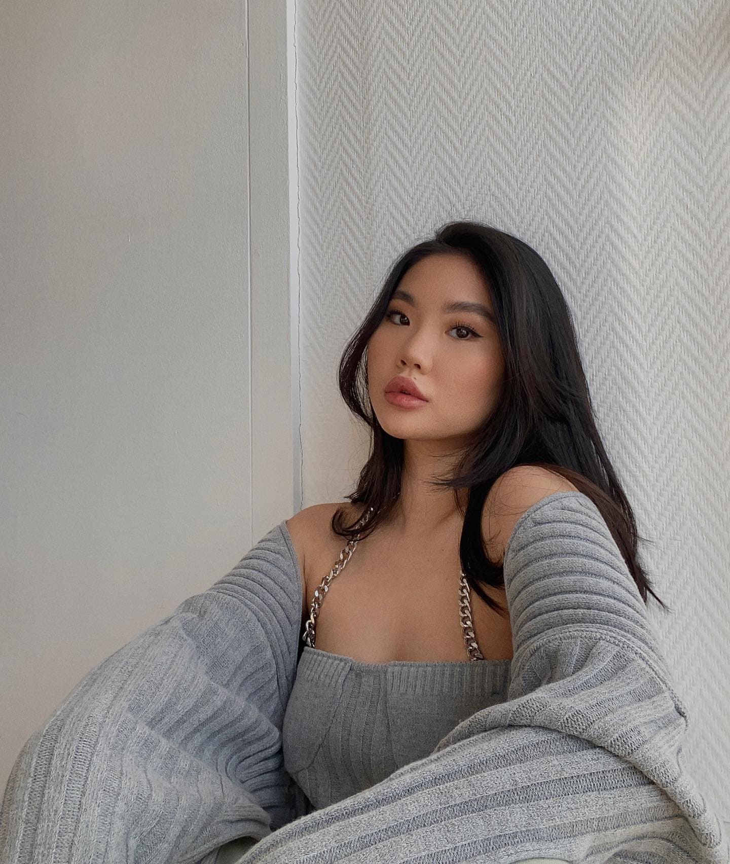 Sitting against a textured white wall in a grey ribbed knit top with chain straps and an off-shoulder cardigan.