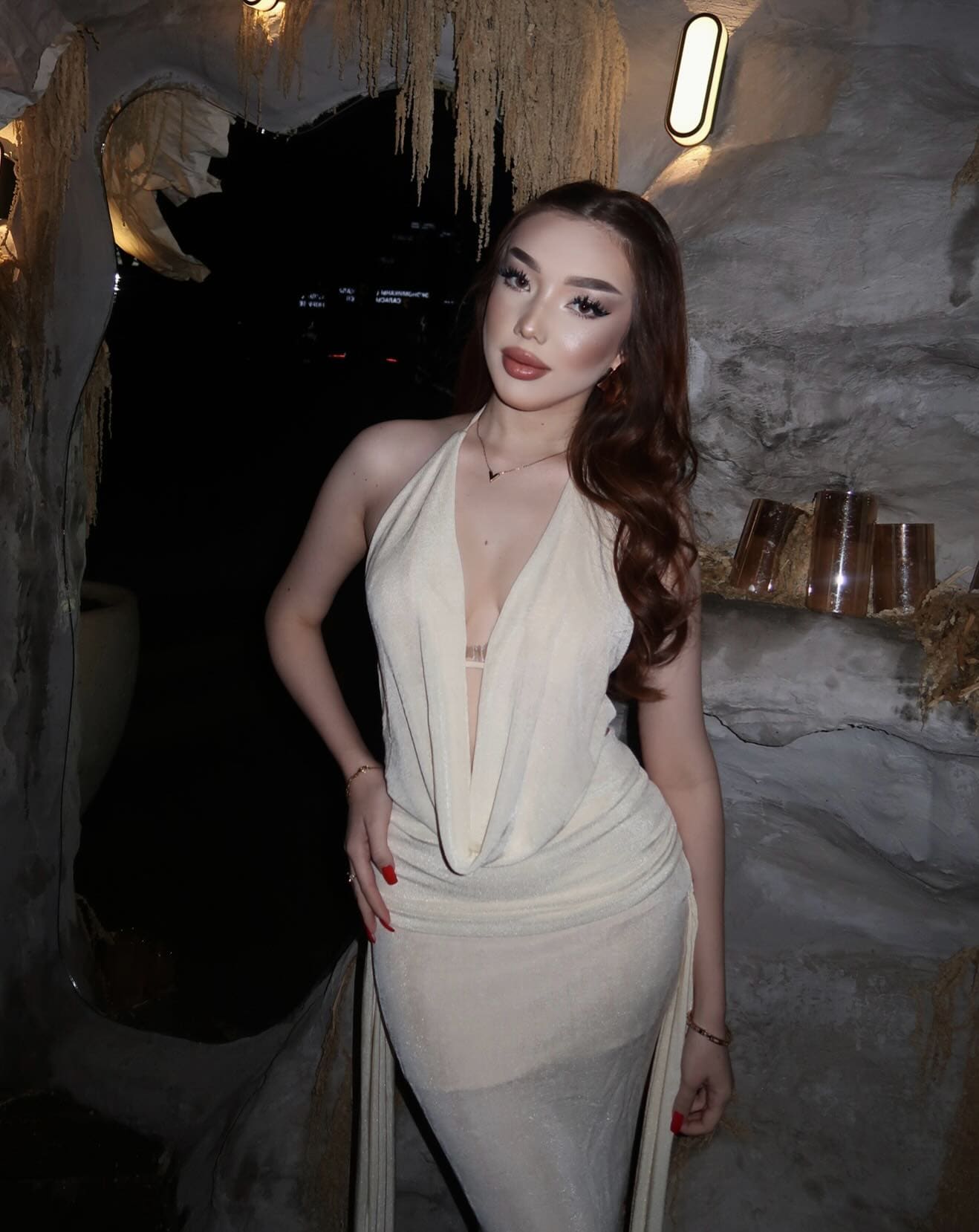 A woman stands in a dim cave-like interior wearing a cream halter dress with a plunging neckline, resting one hand on her hip.