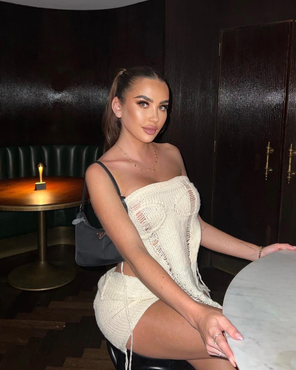 A woman sits on a stool in a dim room wearing a cream open-knit outfit and carrying a small dark handbag.