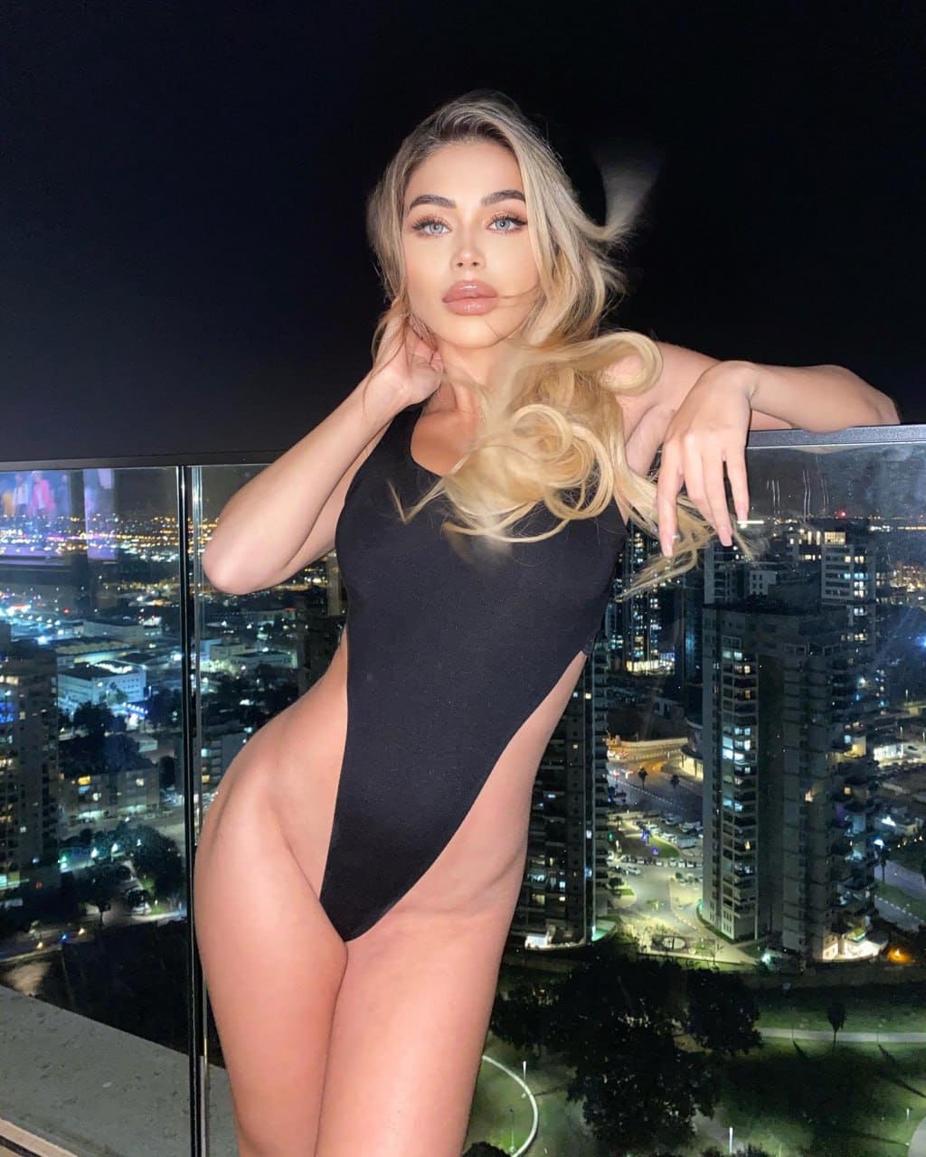 A woman stands on a rooftop at night in a black sleeveless bodysuit, resting one hand on a glass railing with a city skyline behind.