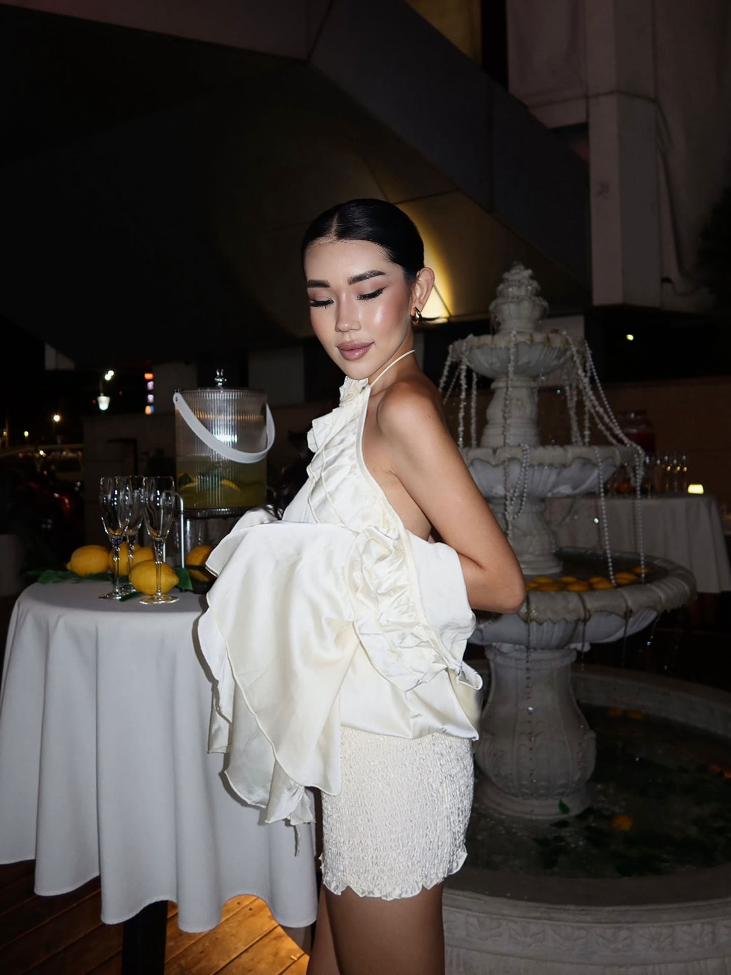 A woman in profile wears a white ruffled halter top and short white skirt, posing by champagne flutes and lemons near a stone fountain.