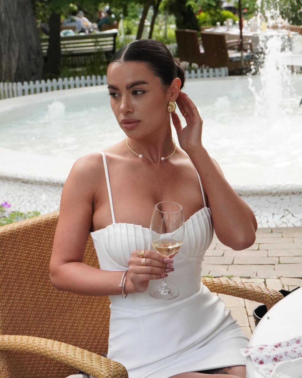 Emily sits in a woven chair outdoors, wearing a white strapless dress and pearl necklace, holding a glass of white wine near a fountain.