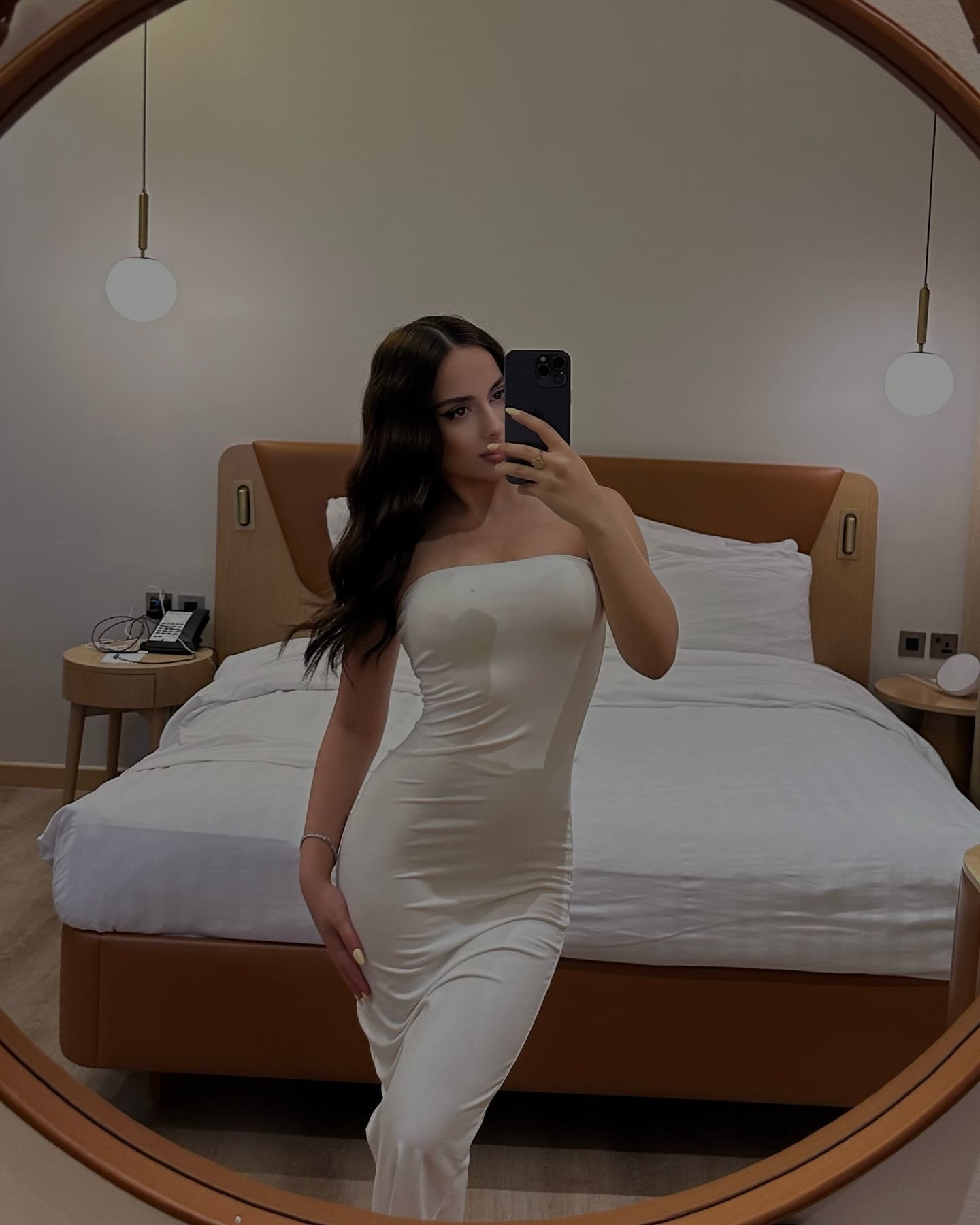 A woman in a white strapless dress takes a mirror selfie in a bedroom, with a bed and two pendant lights visible behind her.