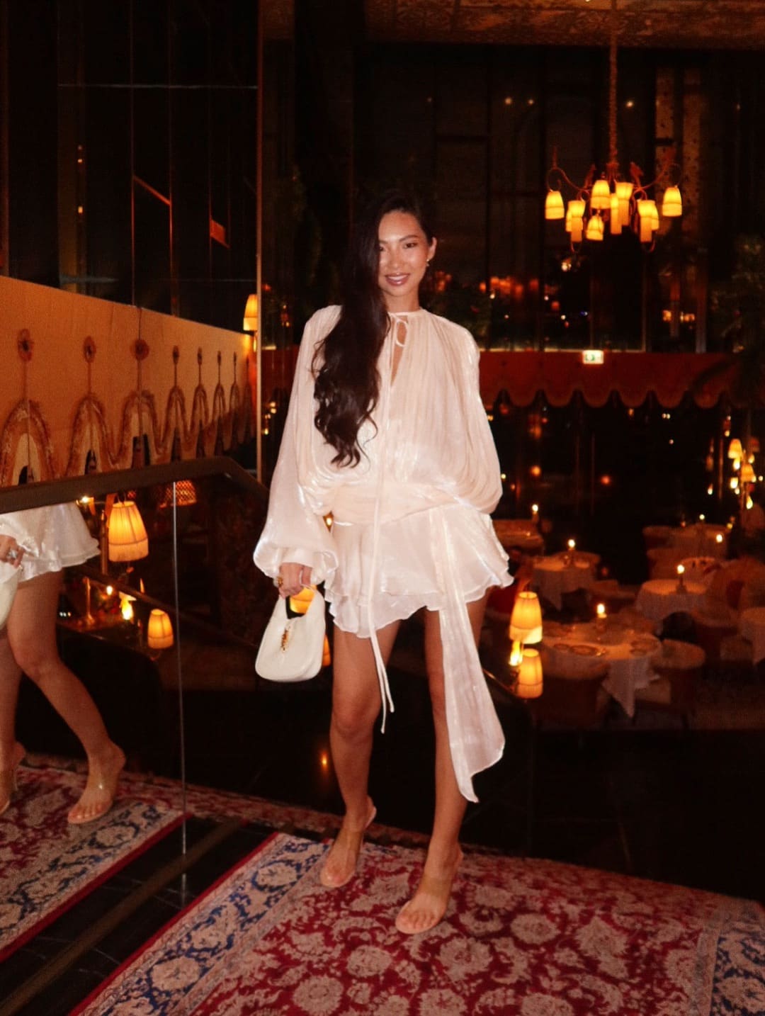 Standing on a rug in a dim restaurant, she wears a sheer white mini dress with a slit, holding a white handbag.