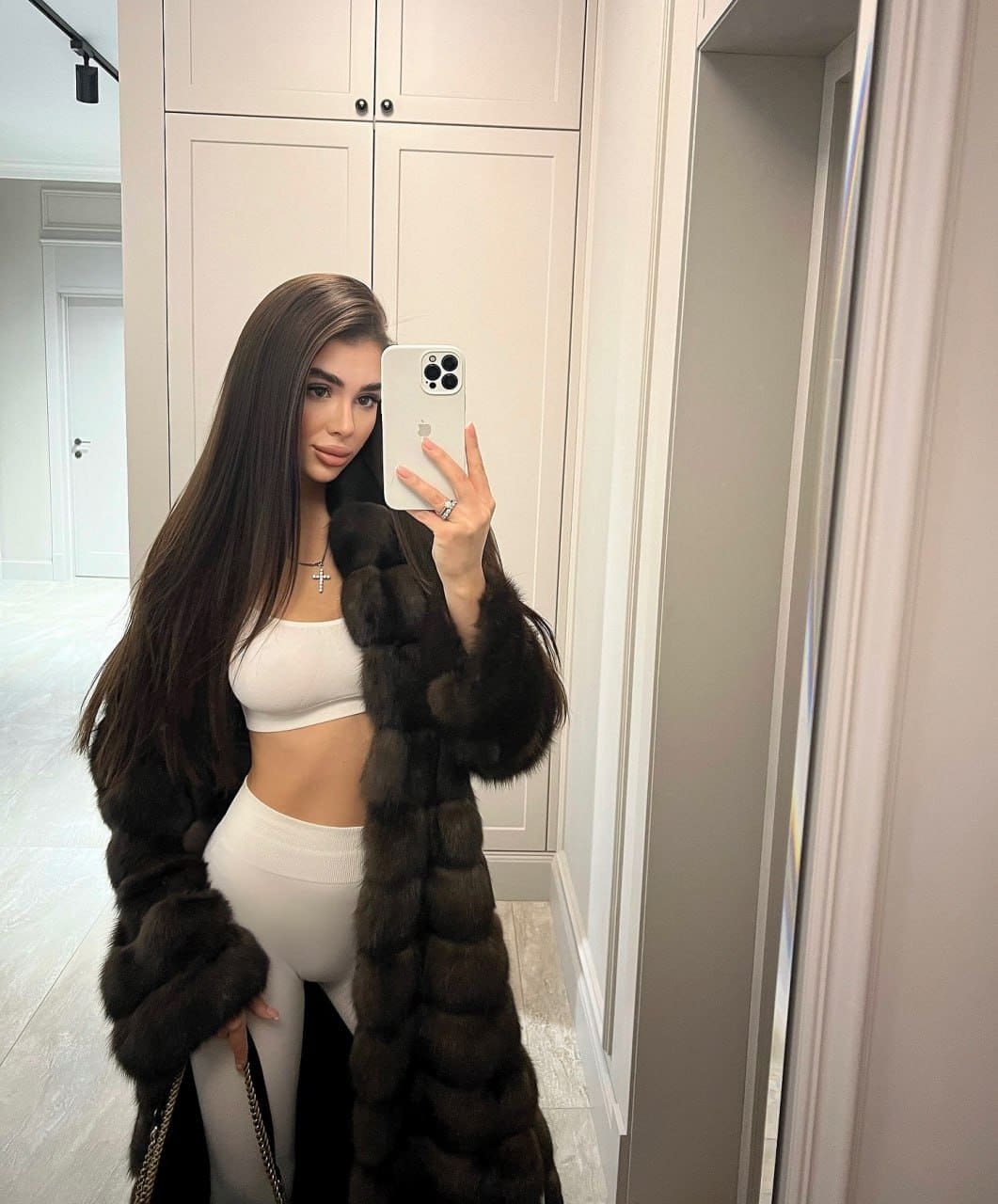A woman takes a mirror selfie in a white room, wearing a white crop top, leggings, and an open dark fur coat.