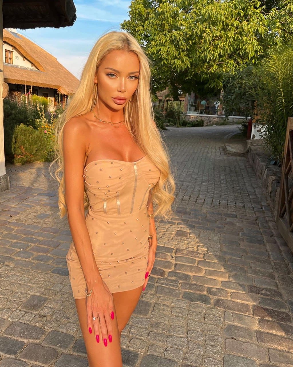 Anya stands on a cobblestone path in a strapless beige dress, hand on thigh, before a thatched building.