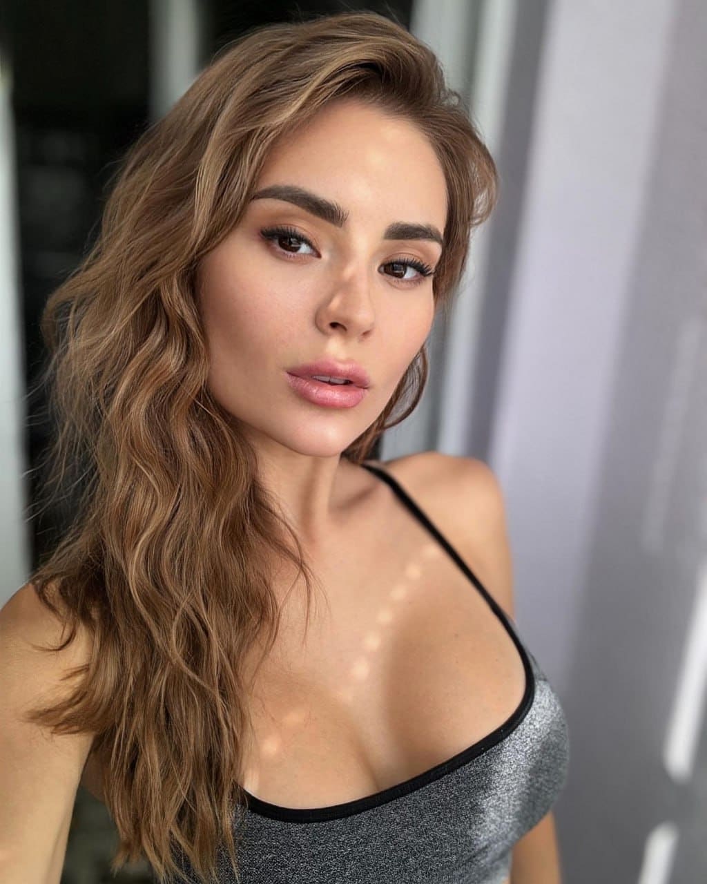 Fiona with long wavy light-brown hair wears a silver glittery top with thin straps, posing against a softly lit gray indoor background.
