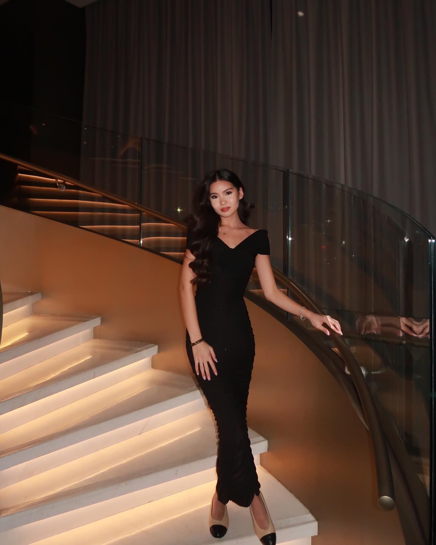 A woman in a fitted black V-neck dress stands on a curved white staircase, resting her hand on a glass railing.