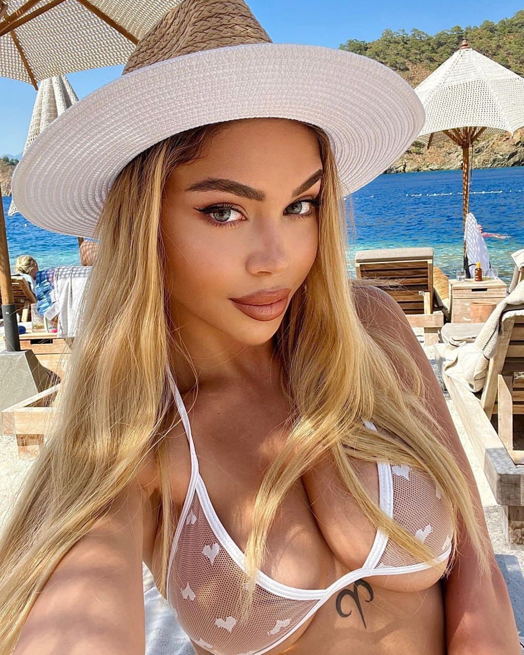 Natalia takes a beachside selfie wearing a wide-brimmed straw hat and a sheer white bikini top with heart patterns.