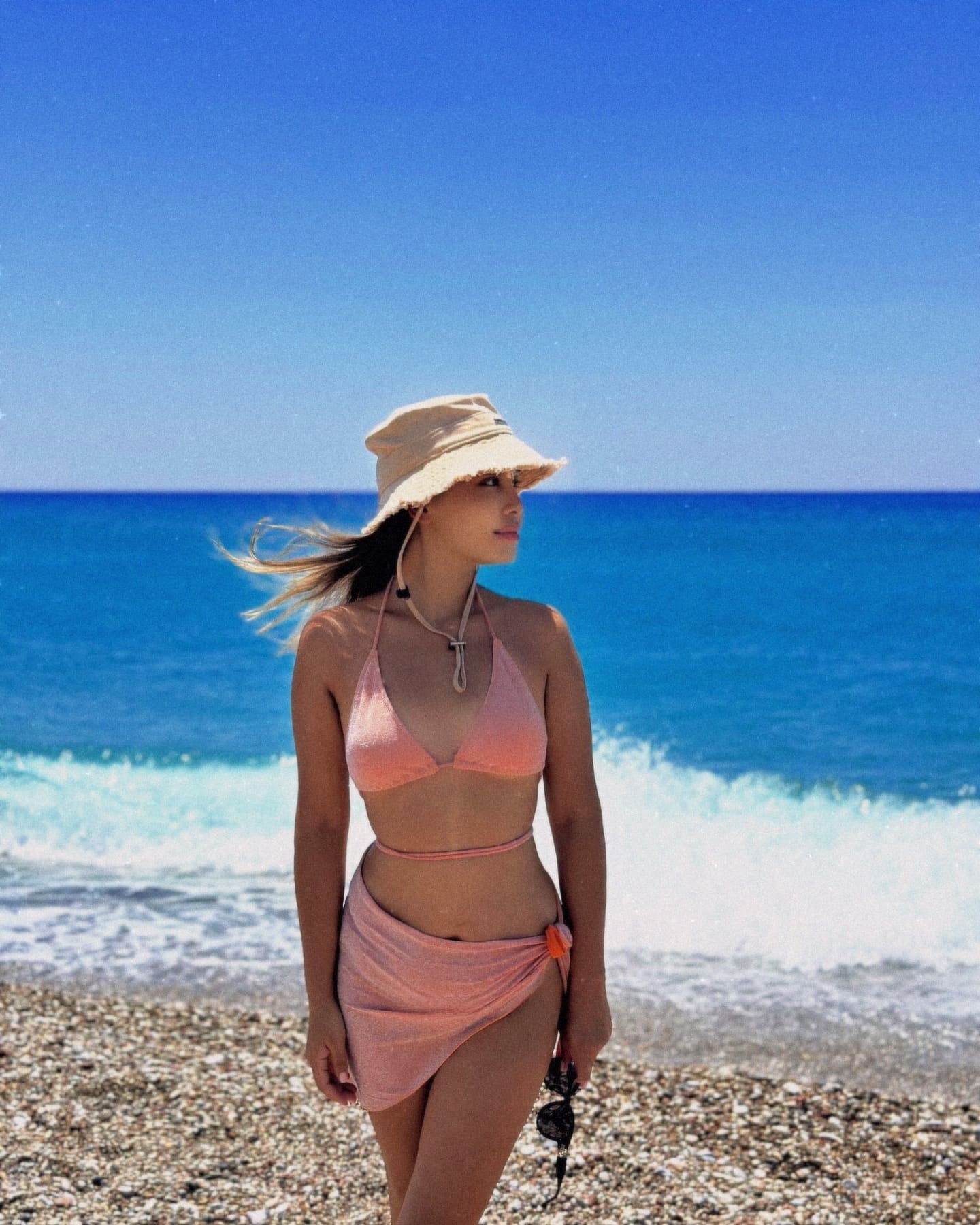 A woman stands on a pebble beach wearing a pink bikini top, wrap skirt, and beige bucket hat, holding sunglasses against a blue ocean backdrop.