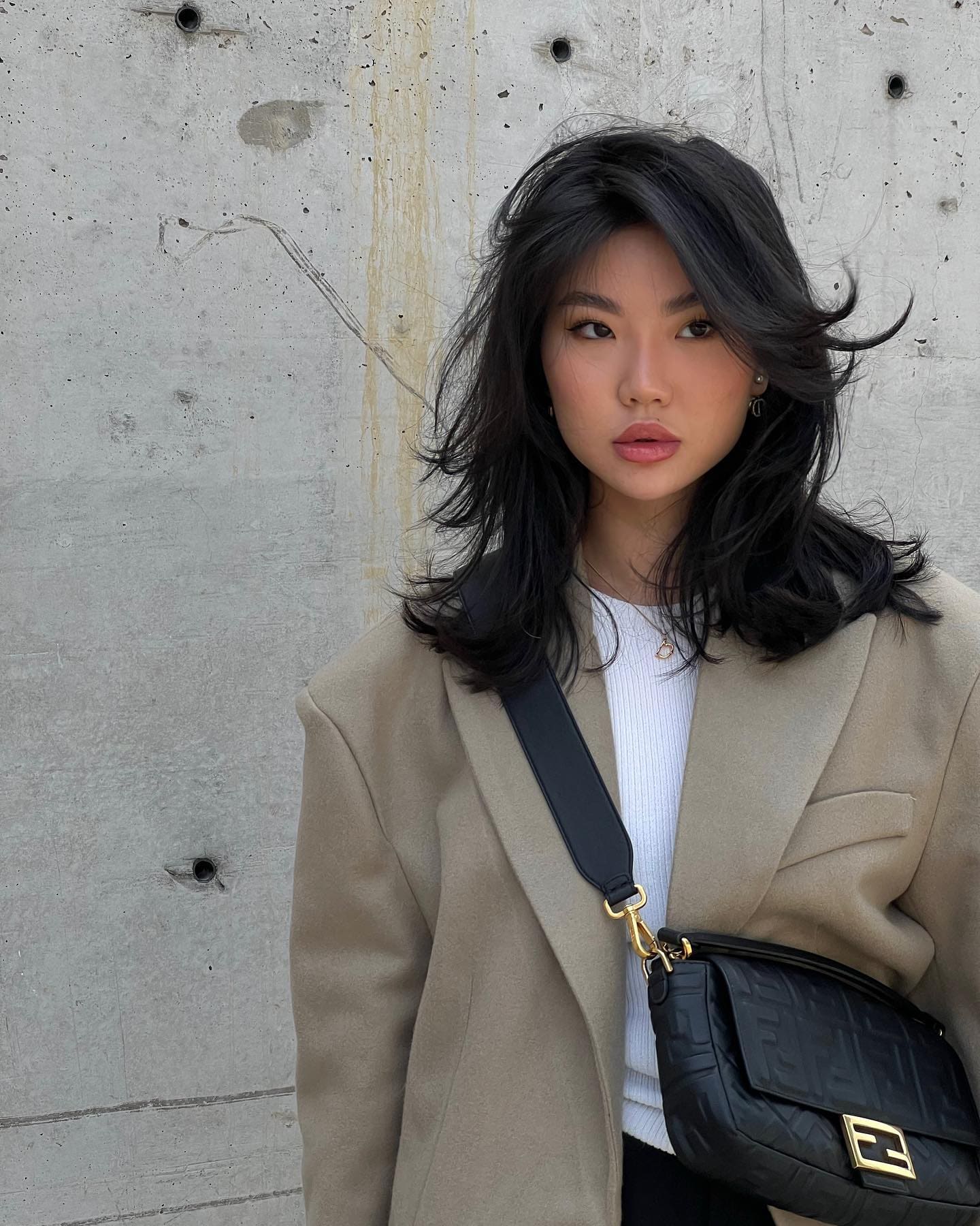 Dinara stands against a gray concrete wall in a beige blazer, white ribbed top, and black quilted crossbody bag.