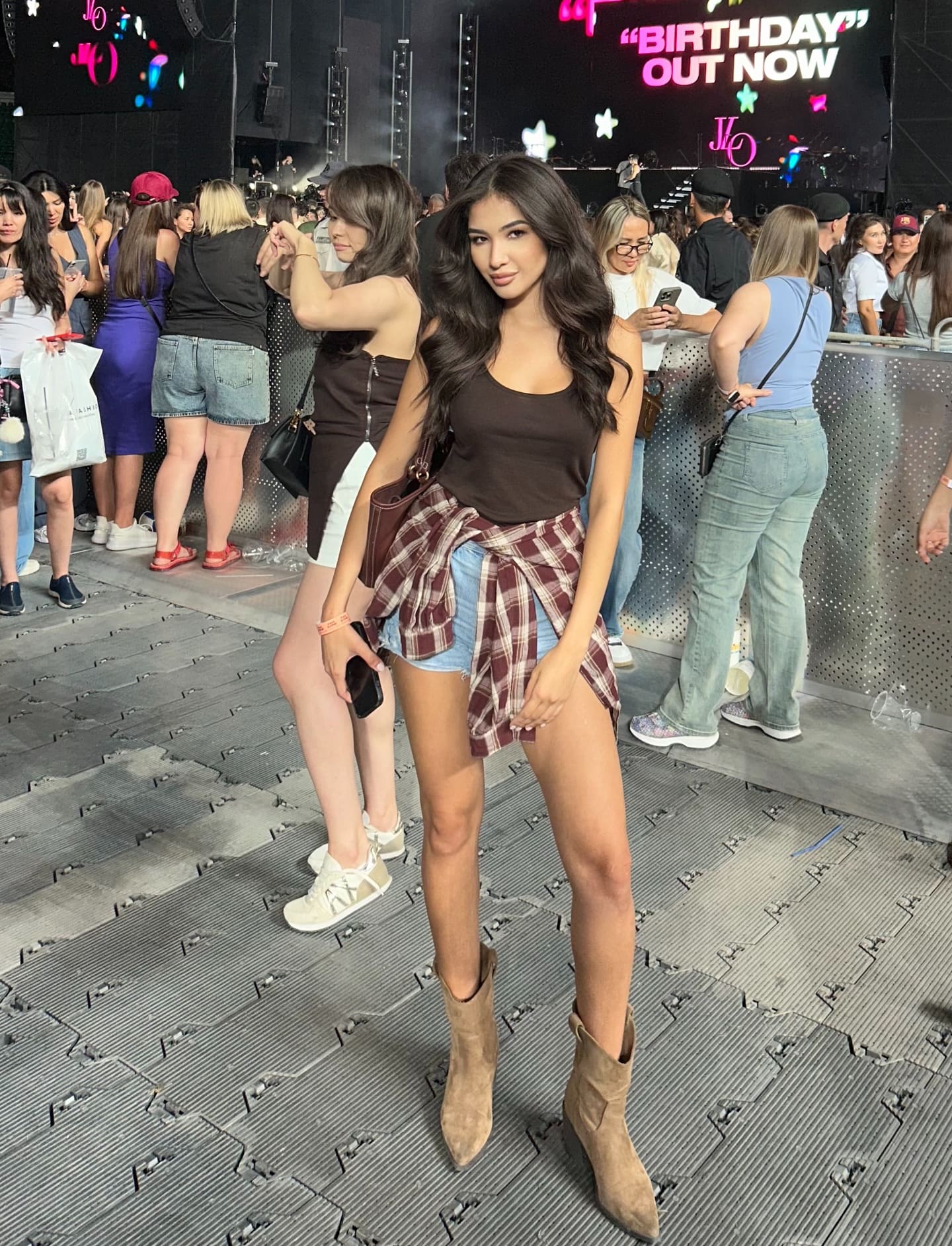 A woman in a brown tank top, denim shorts, and a plaid shirt tied at the waist stands outdoors holding a phone, with a stage screen behind her.