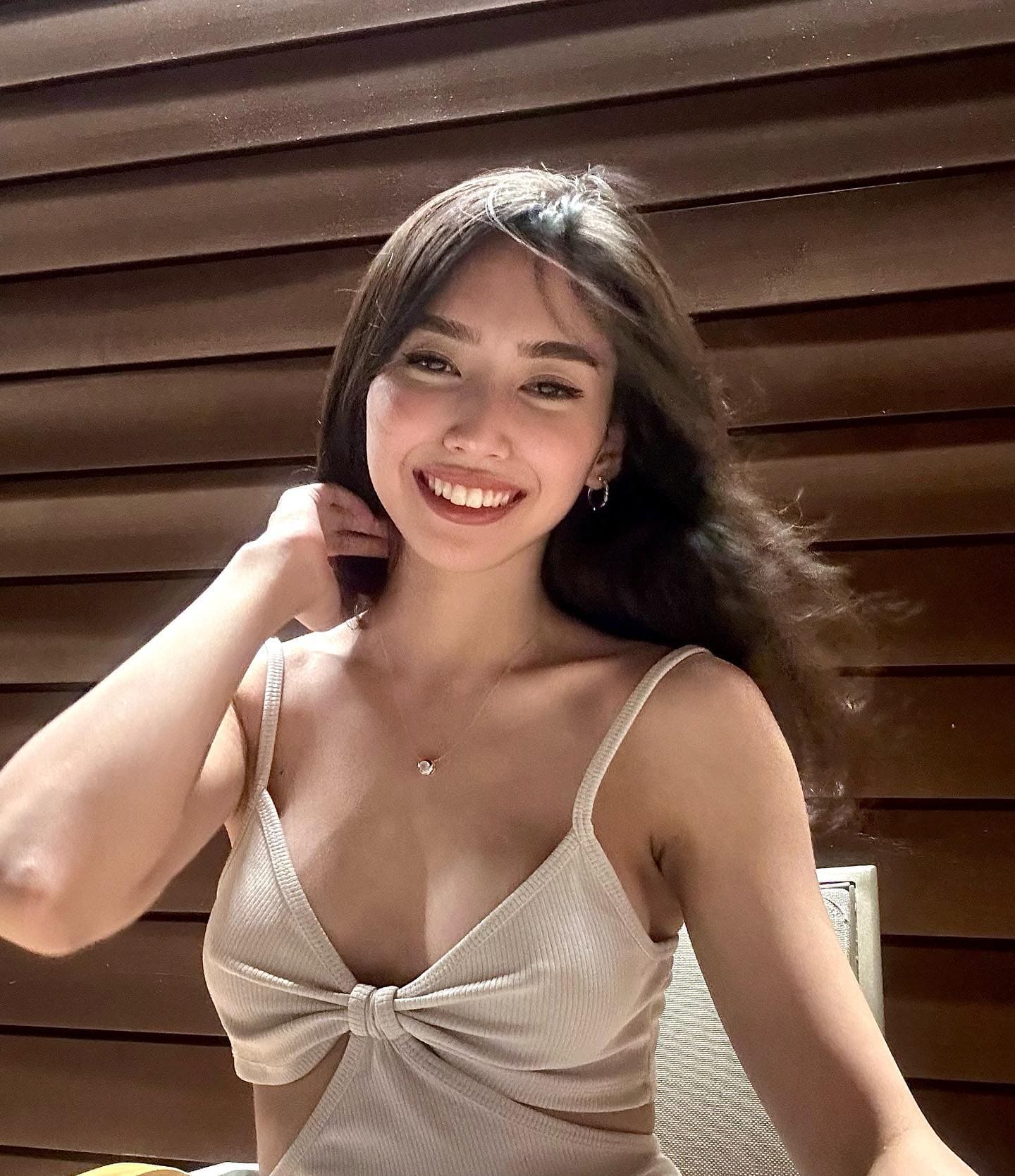 Elvina smiles in a beige ribbed top with thin straps and a deep V-neck, posing by a slatted wooden wall with one hand near her neck.